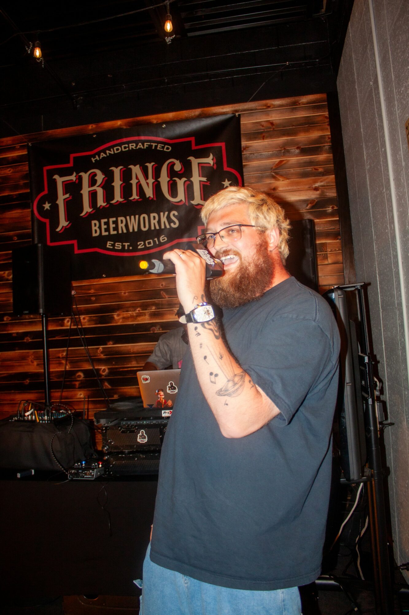 Person with glasses and a beard singing into a microphone at a bar with a 'Fringe Beerworks' sign behind them.