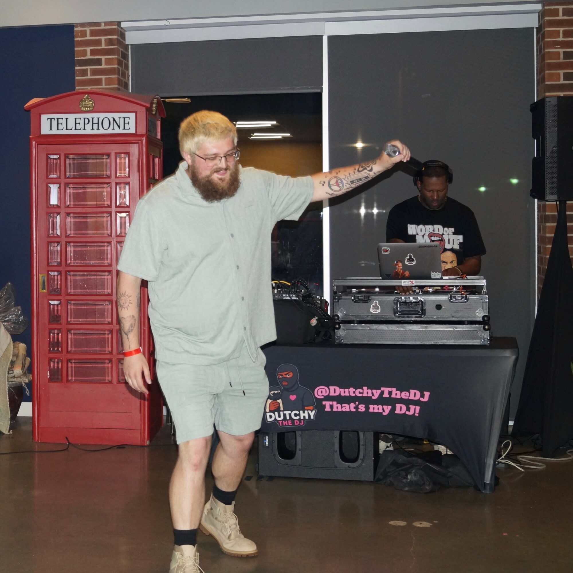 Person dancing with arm raised, DJ in background, red telephone booth, indoor setting, casual attire, smiling, wooden floor.