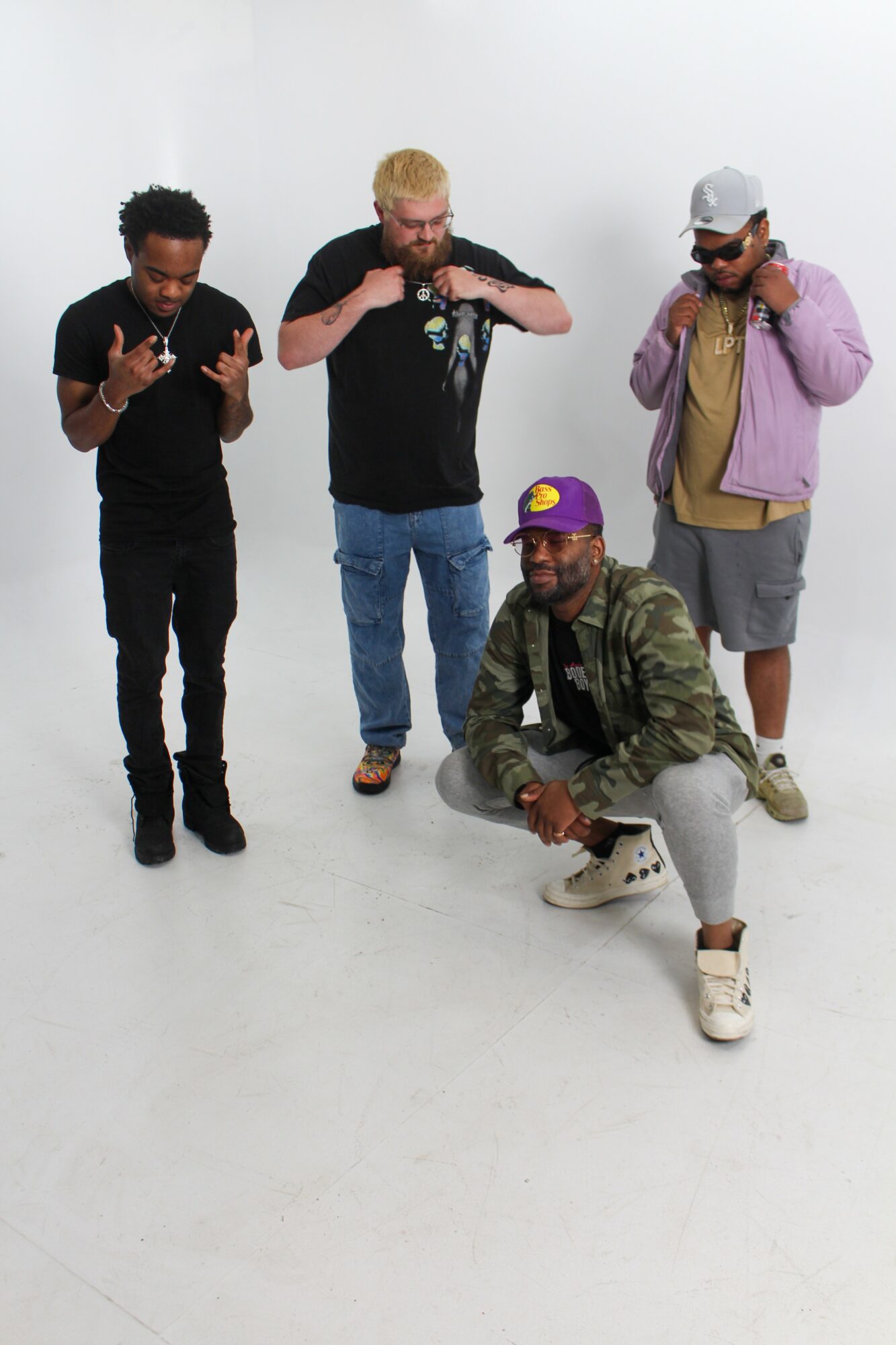 Four men pose together in a white studio, with one crouching and three standing, all making gestures or poses.