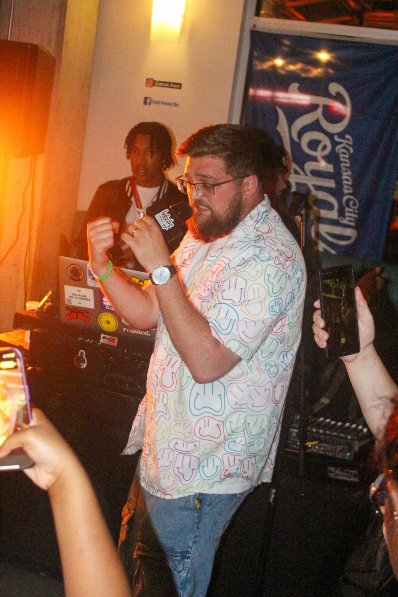 Man with glasses and beard dancing indoors, wearing a patterned shirt and jeans, with a woman in the background and a banner.
