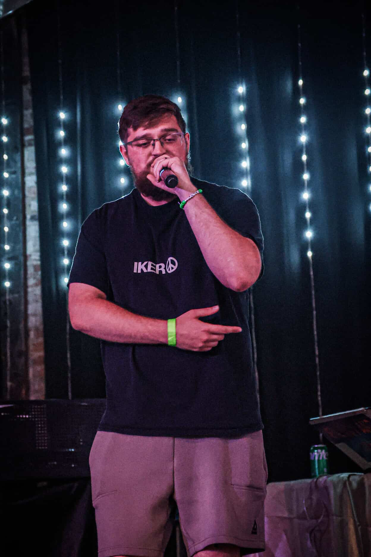 Young man singing into a microphone on stage with vertical string lights in the background.