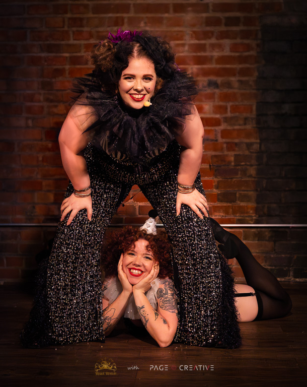 Two women pose playfully against a brick wall, one standing with hands on knees and the other lying on the floor, smiling.