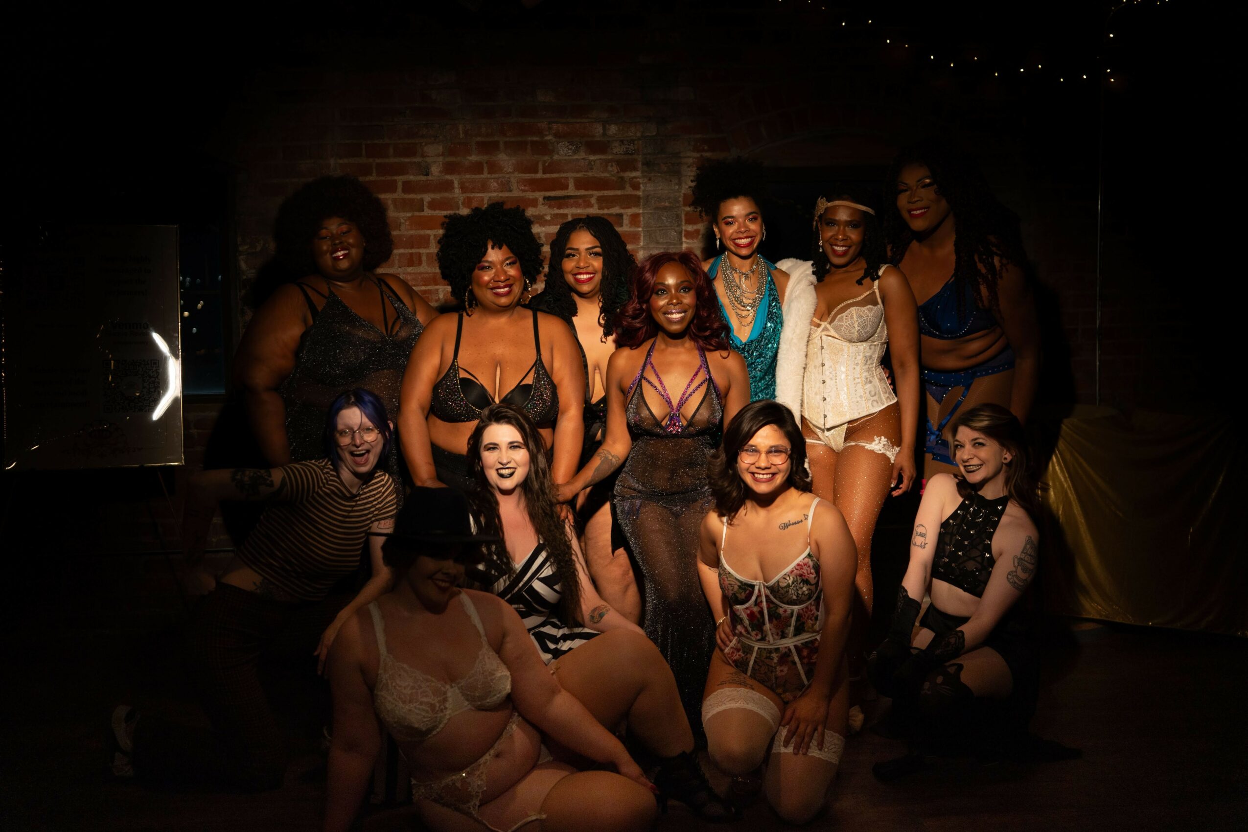 Group of women posing together on stage with a brick wall background, some wearing costumes and accessories.