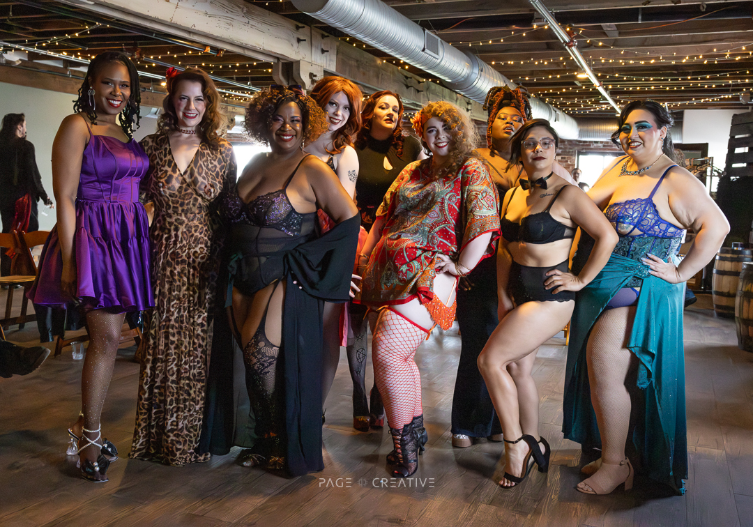 Group of nine women in colorful outfits posing indoors with string lights and wooden flooring.
