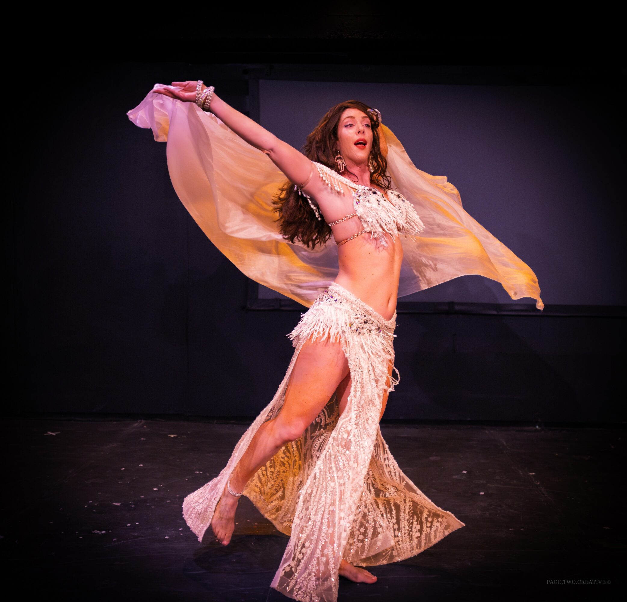 A woman in a flowing costume performs a dance on stage with arms raised and fabric flowing around her.