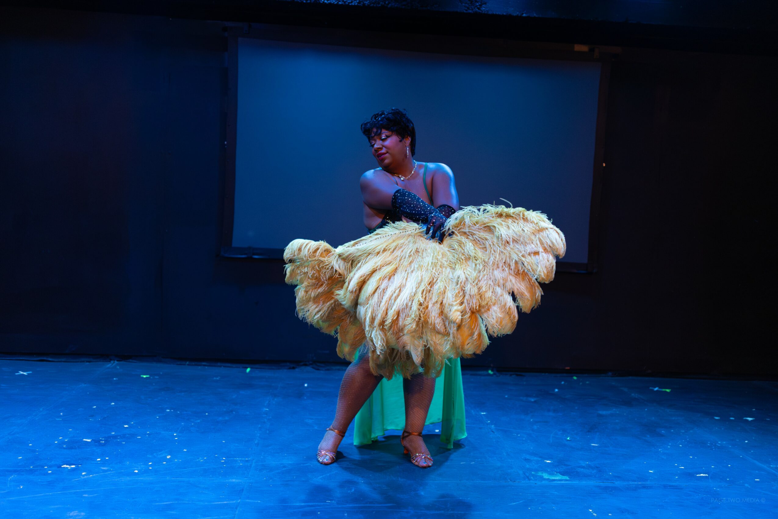 Person standing behind a large, fluffy, yellowish-brown costume or prop, on a stage with a blue background.