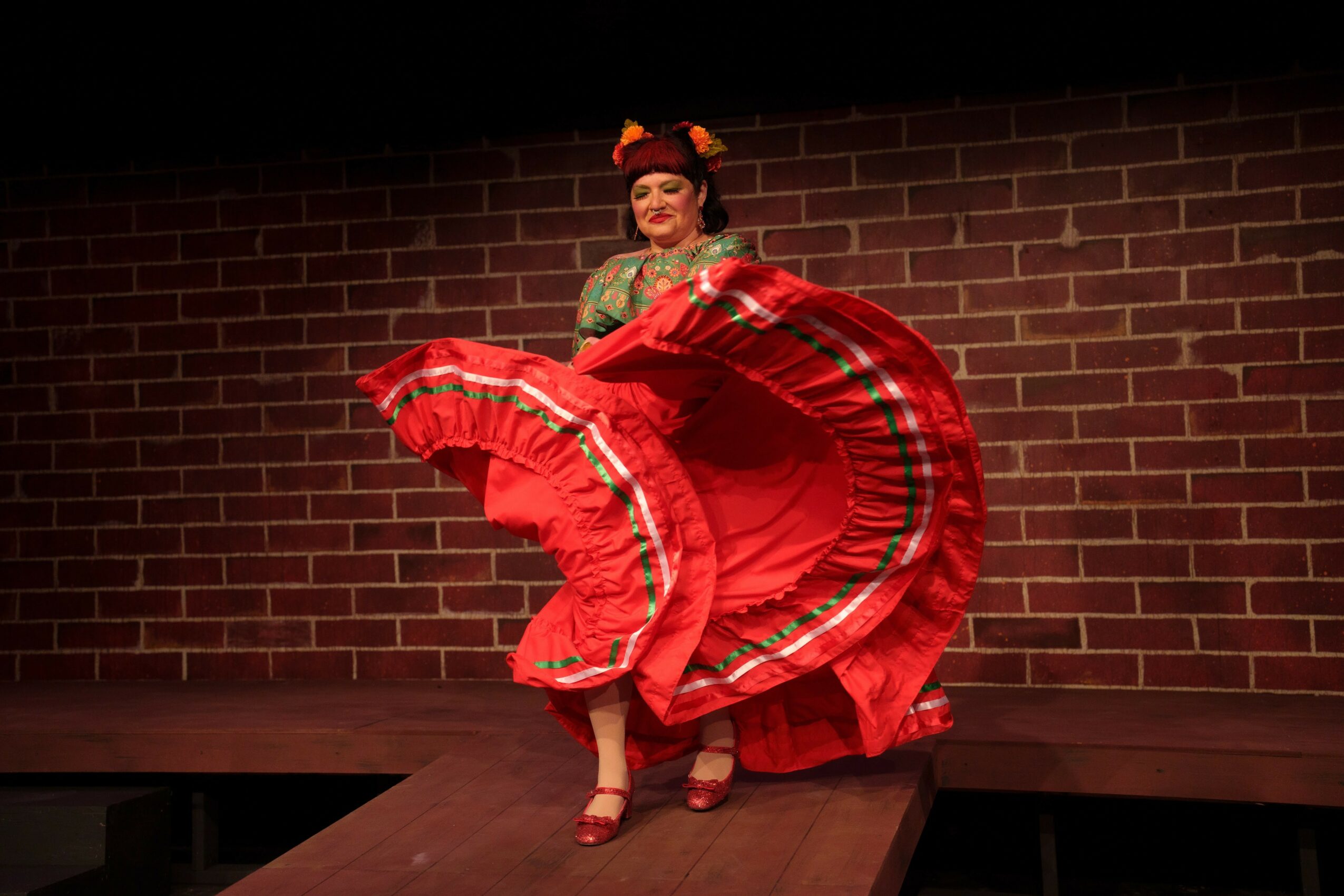 Person in traditional costume holding a large red skirt, standing on a stage with a brick wall background.