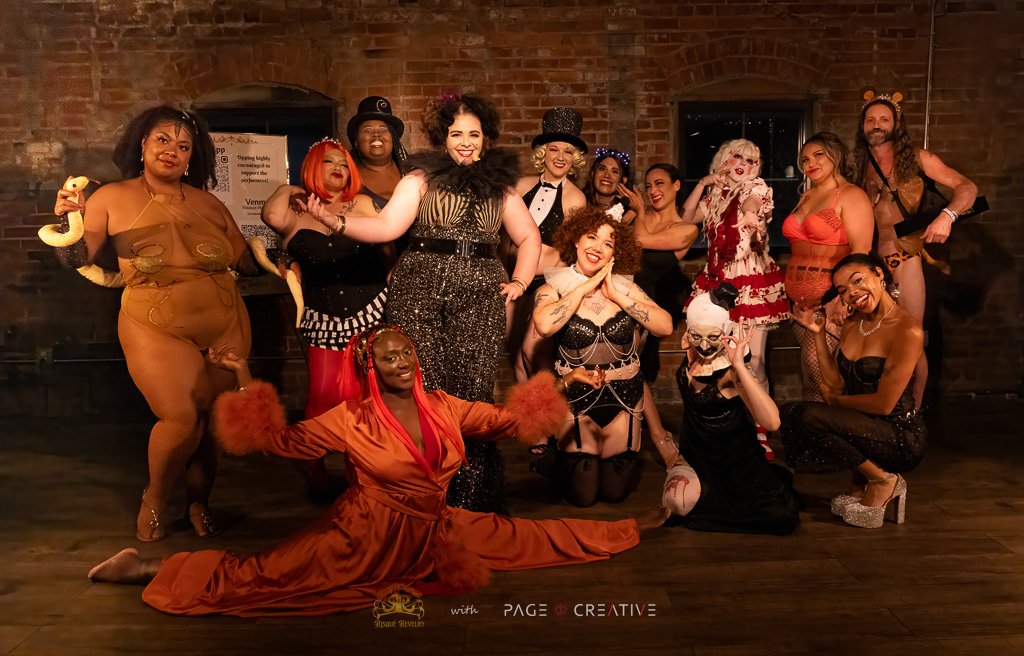 Group of people in festive costumes posing indoors with brick wall background, some holding drinks, celebrating together.