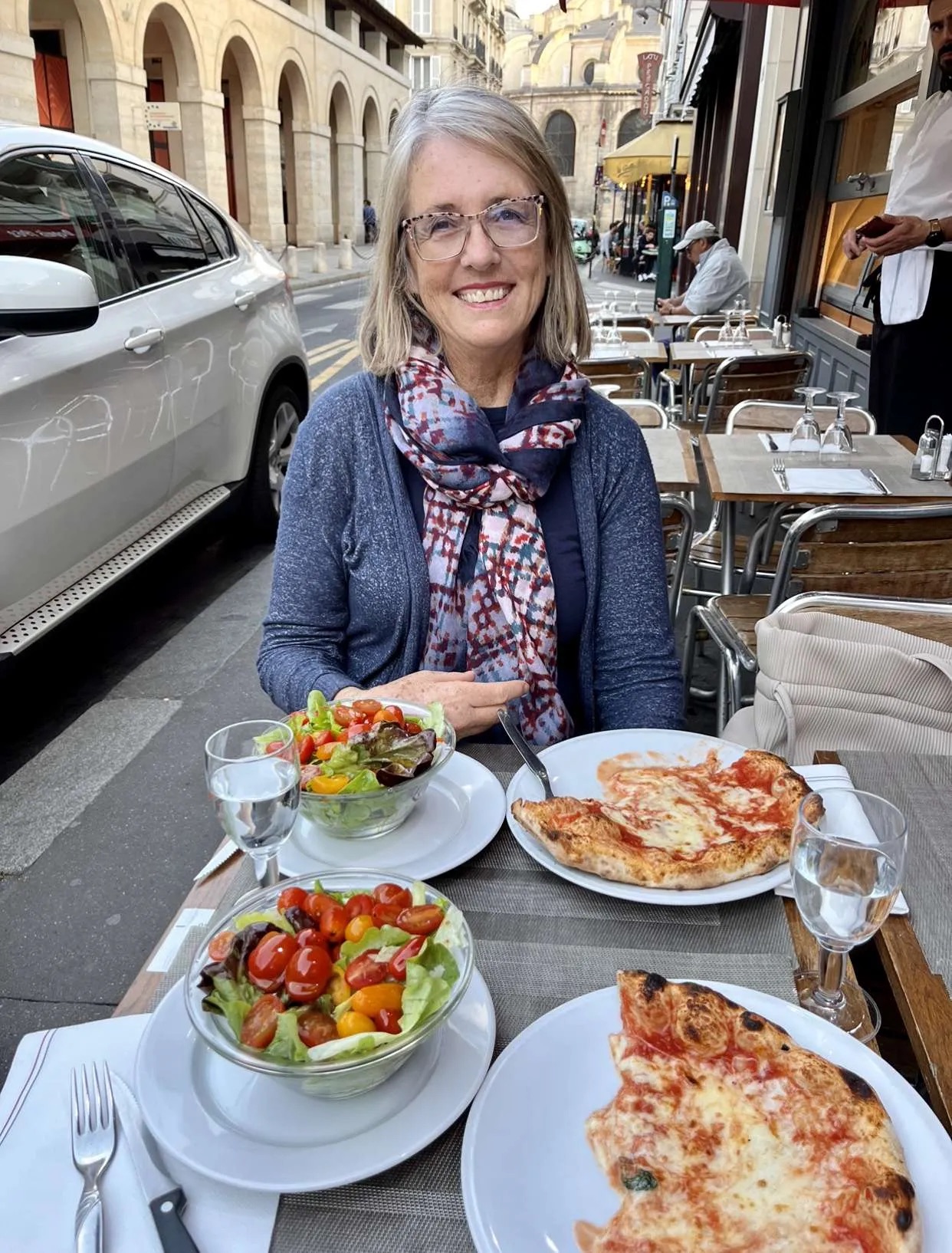 Smiling woman with glasses and a scarf sitting at an outdoor cafe table with salads and pizza, city street background.