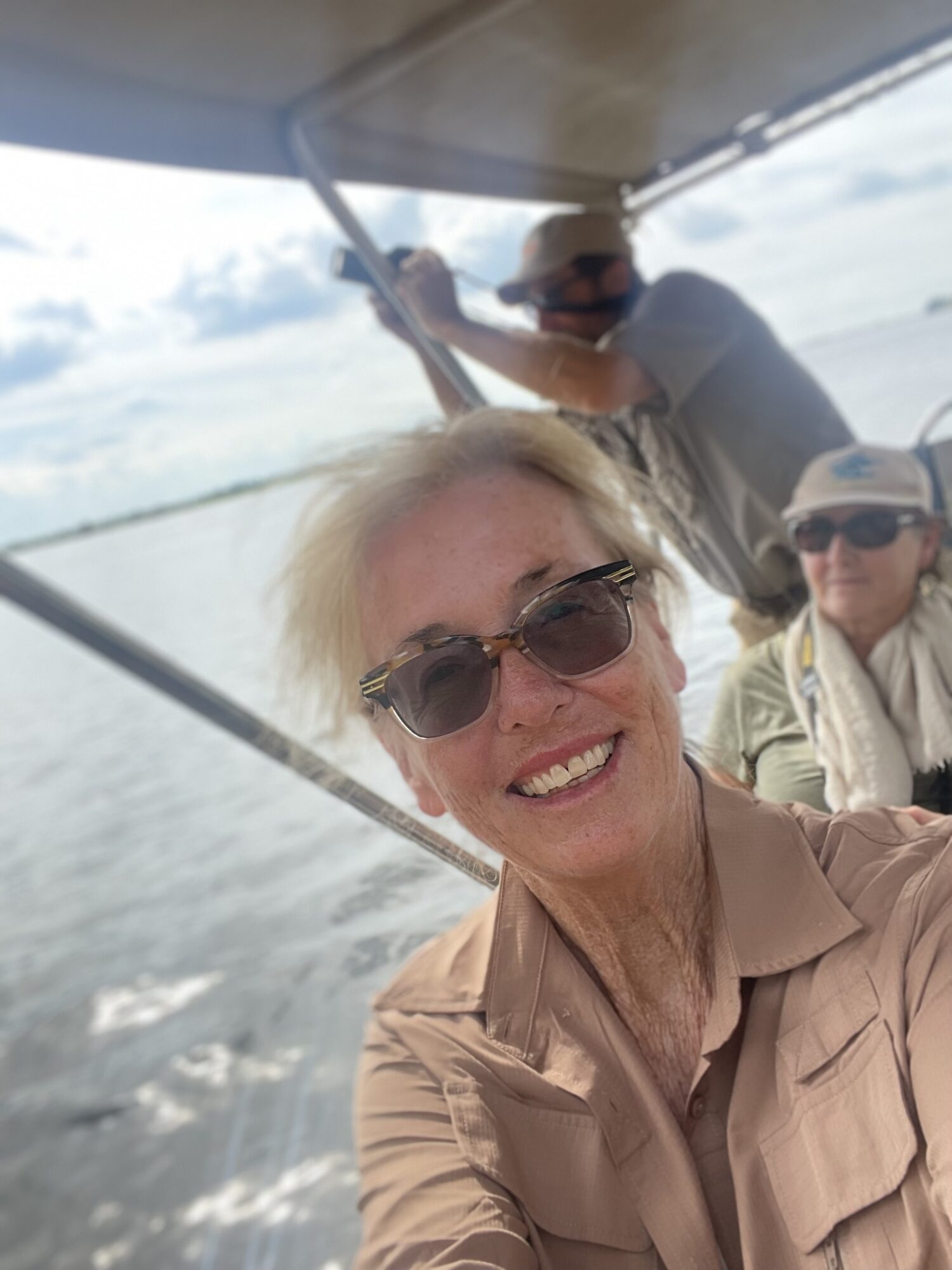 Smiling woman wearing sunglasses on boat with two people in background, water and sky visible.