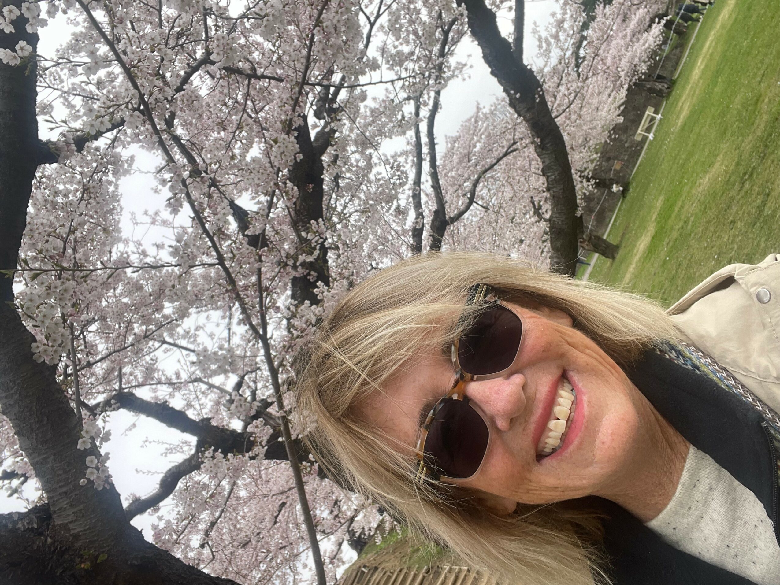 Woman with sunglasses smiling outdoors near blooming cherry blossom trees.