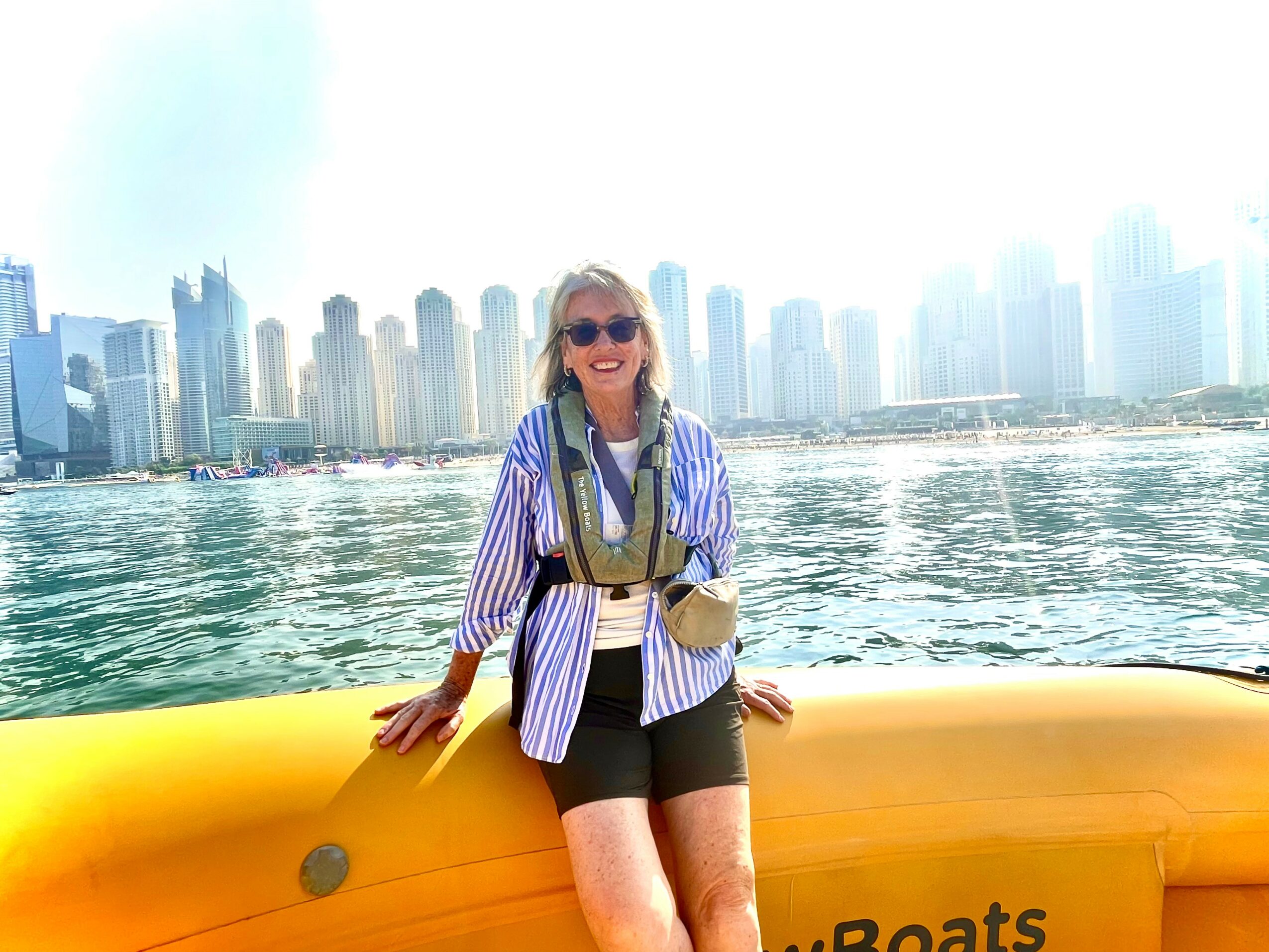 Woman in sunglasses smiling, standing by yellow boat, city skyline and water in background.