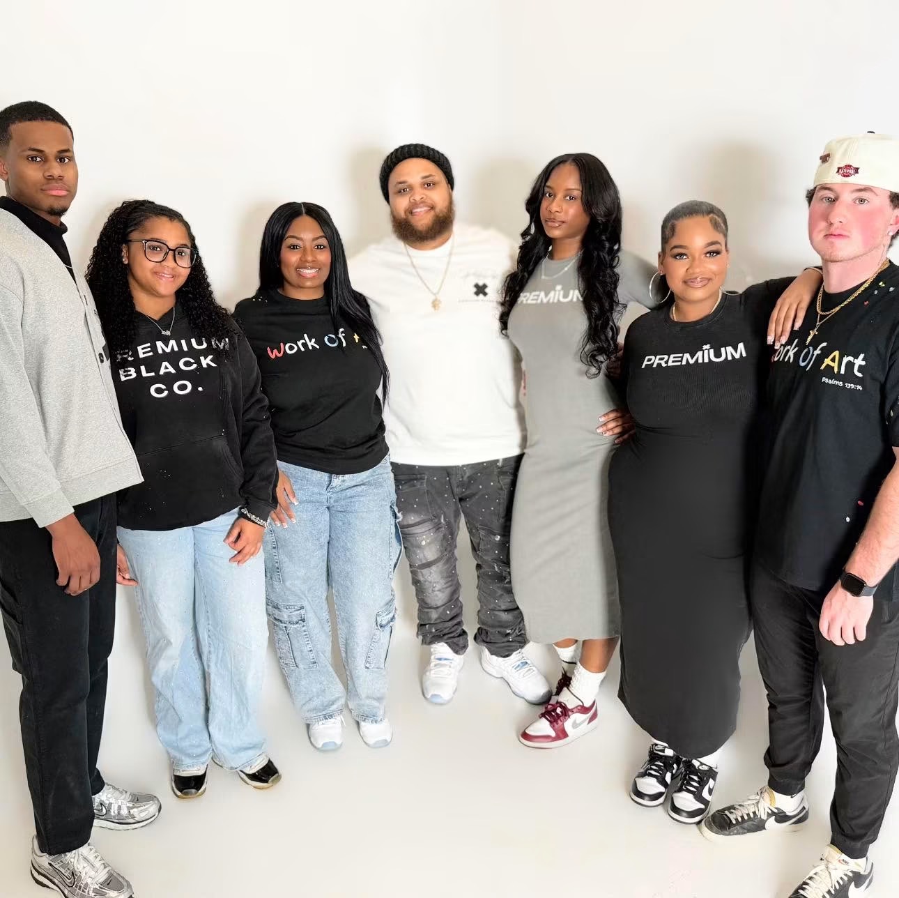 Group of seven diverse people standing against a white wall, smiling, dressed casually, some wearing black clothing, one in a gray dress.