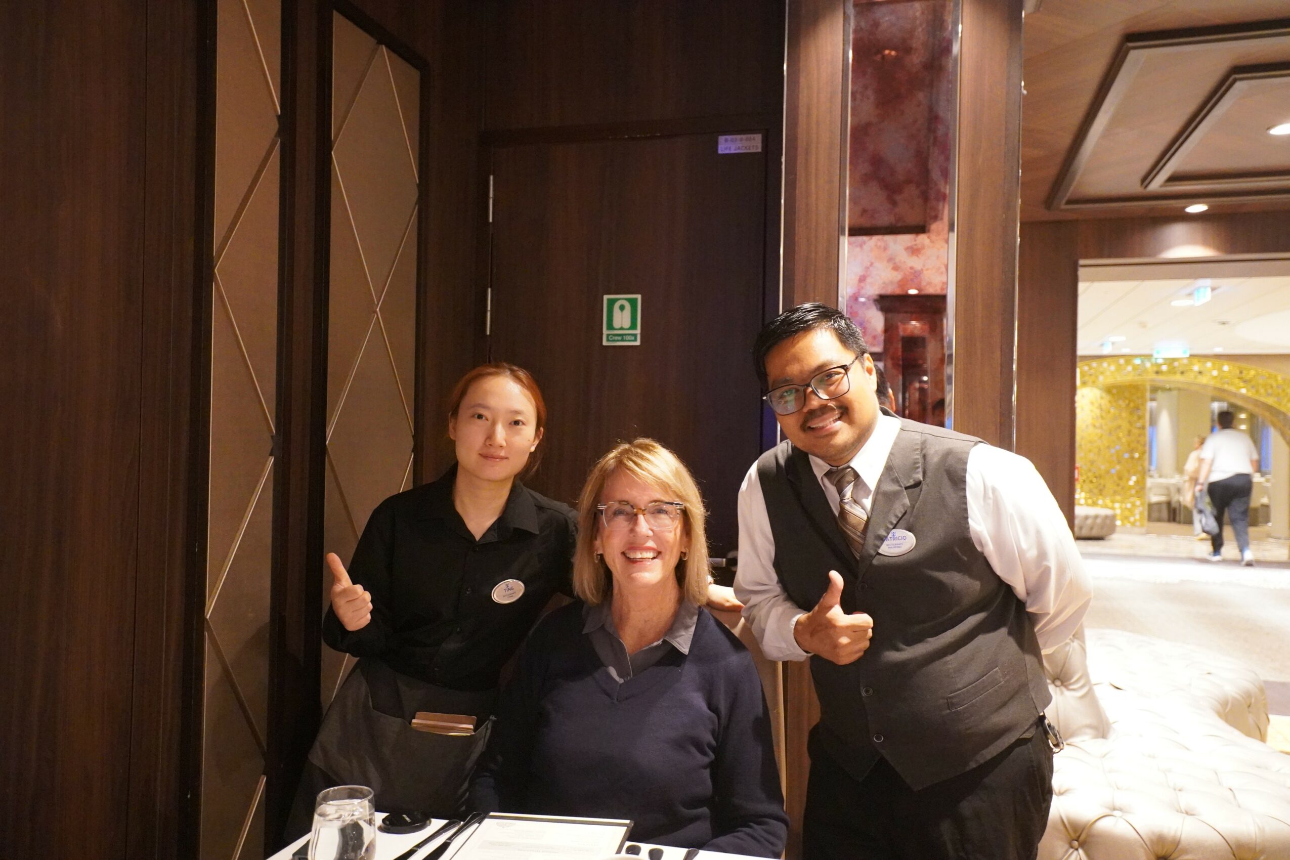 Three people, two women and one man, smiling and giving thumbs-up in a hotel lobby or restaurant, with warm lighting and wooden decor.