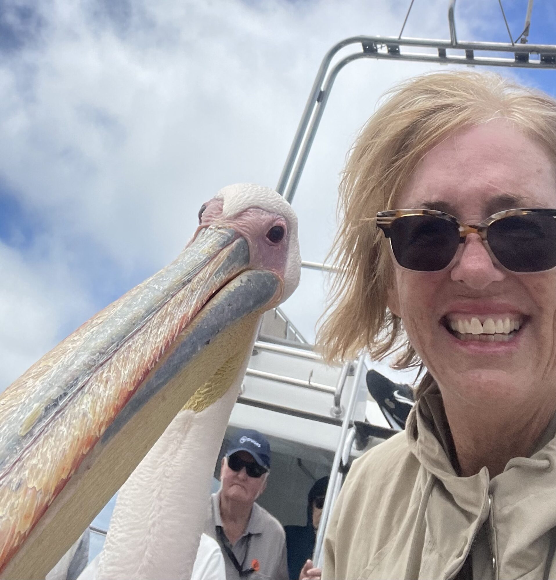 Woman with sunglasses smiling next to a large pelican on a boat, with a man in the background, blue sky and clouds.