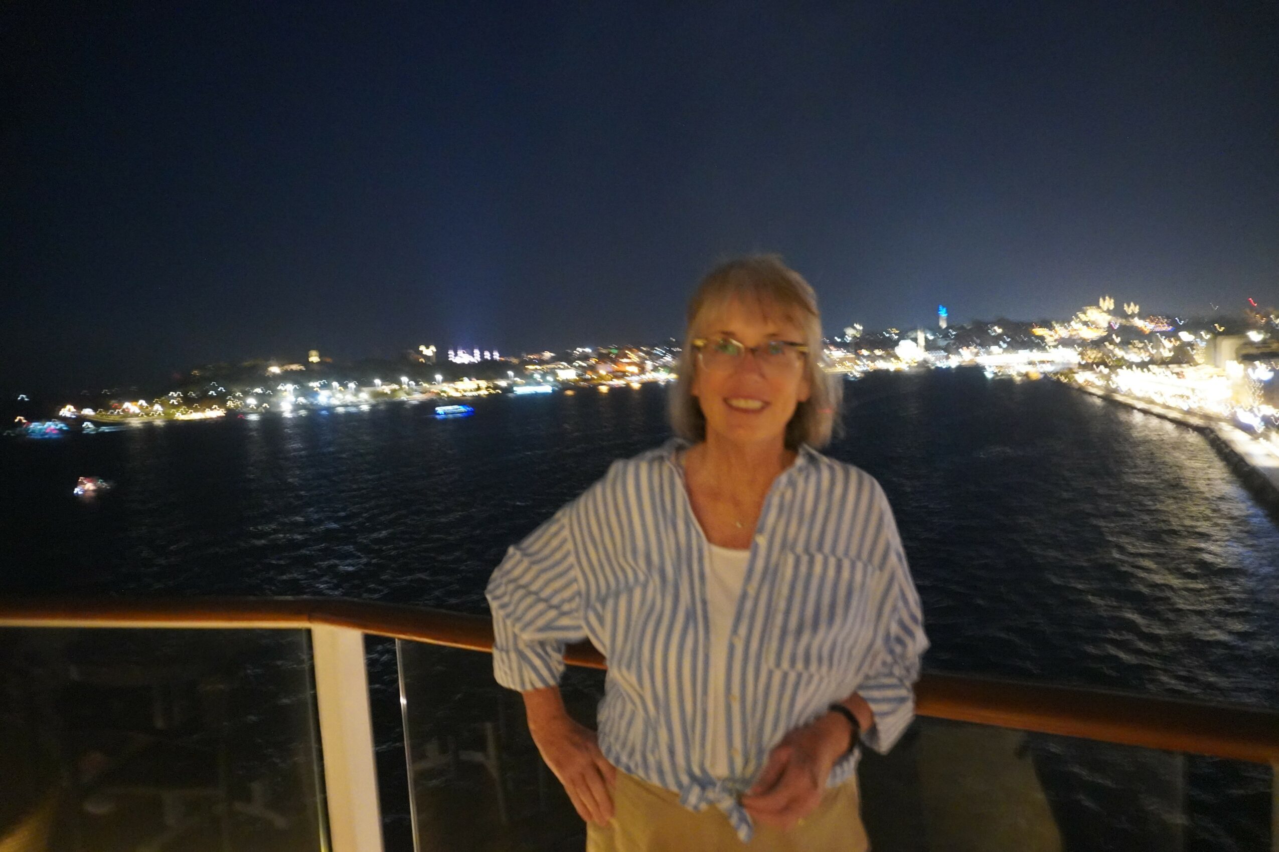 Woman with glasses smiling on a boat at night, city lights in the background, holding a railing.