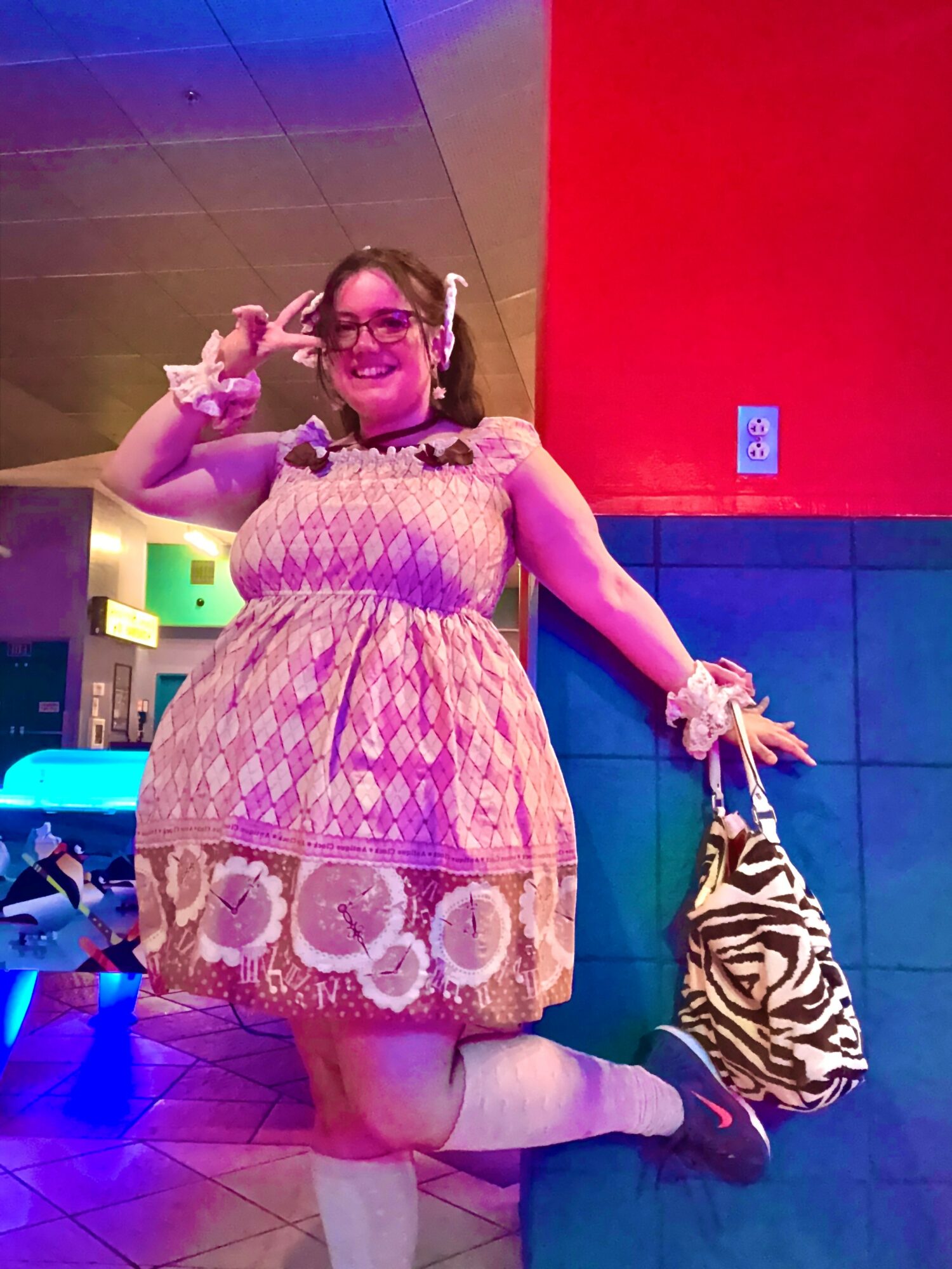 Person with glasses and headphones, smiling, wearing a pink patterned dress, kneeling with a zebra-striped bag, in a colorful indoor setting.