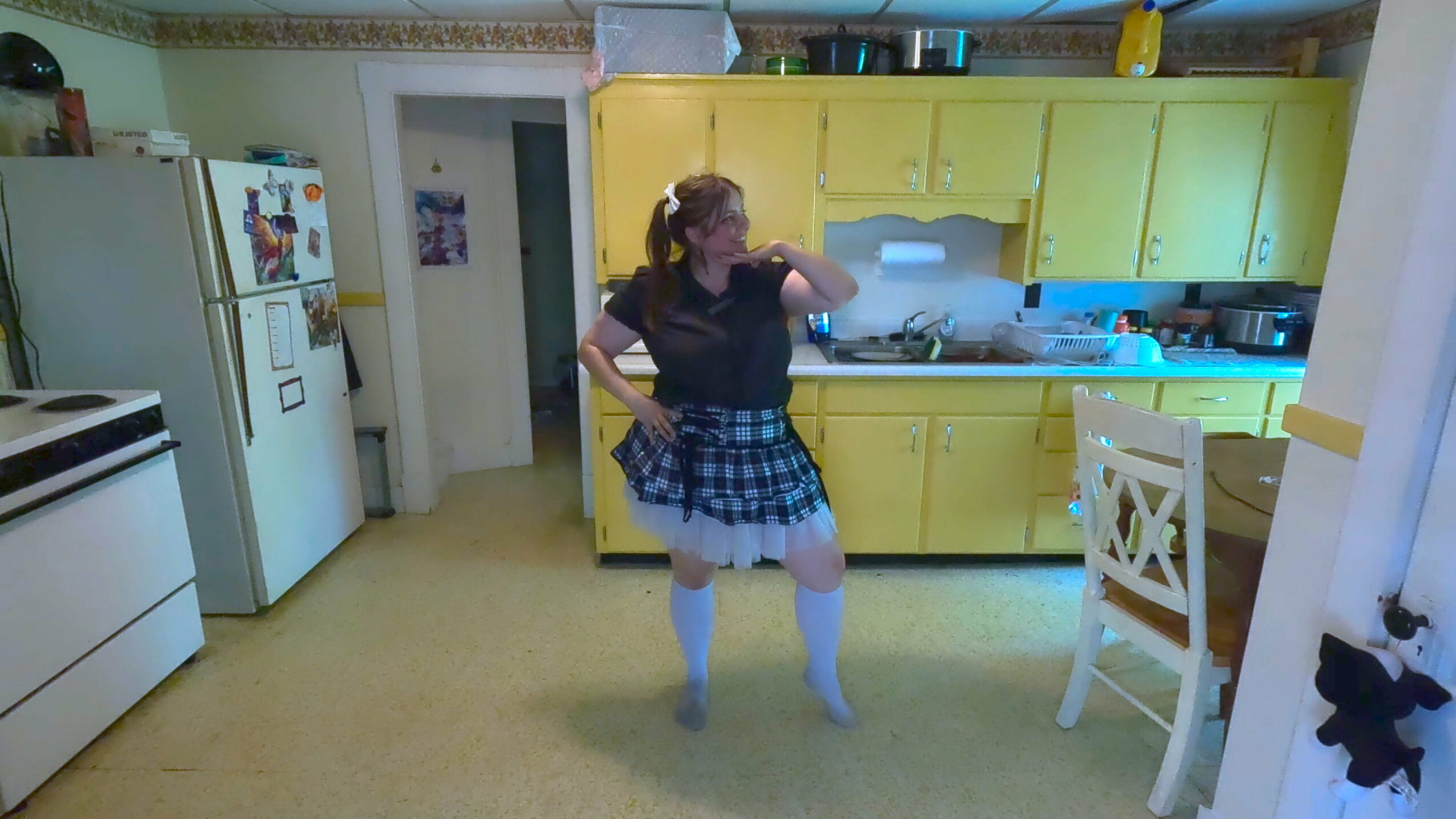 Person dancing in a kitchen with yellow cabinets, wearing a black top, plaid skirt, and white knee-high socks.