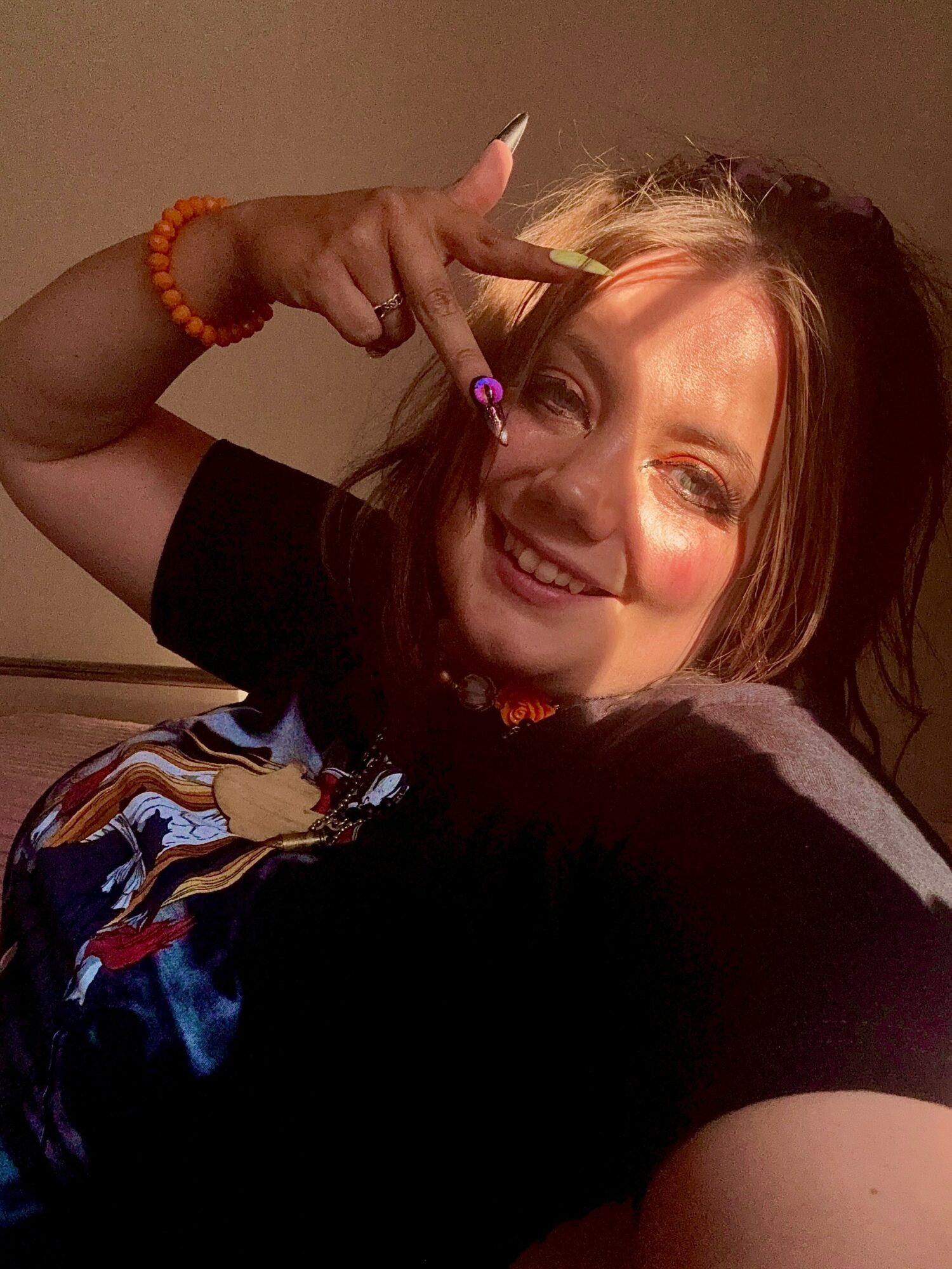 Smiling woman with light brown hair, wearing a black shirt, making a peace sign near her forehead, with a colorful blanket partially visible.
