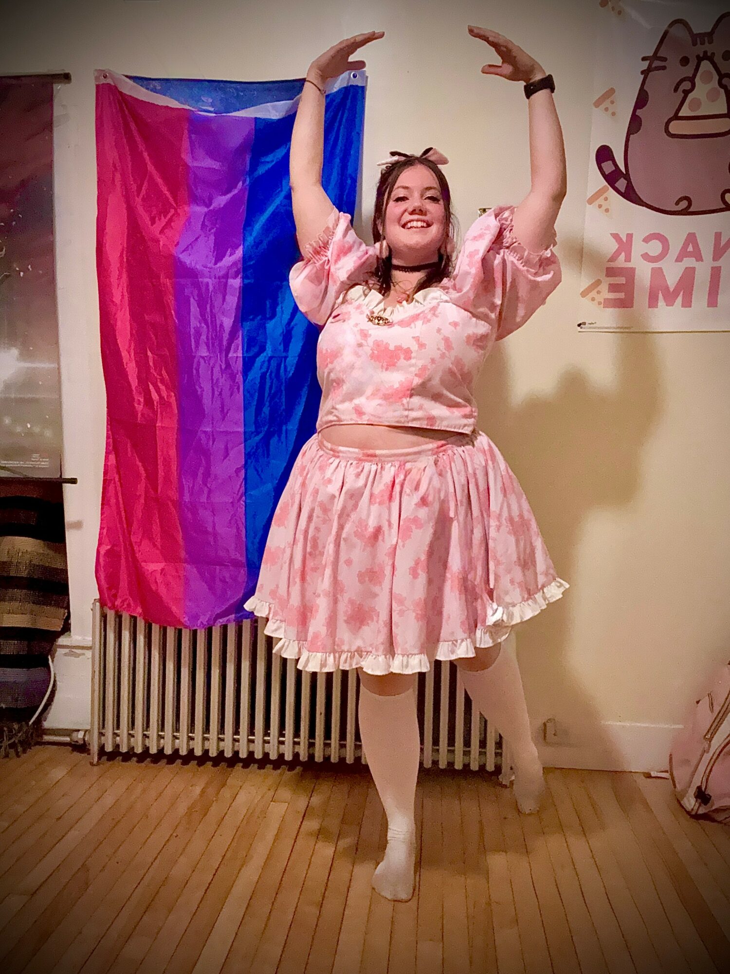 Person dancing with arms raised, wearing pink dress, standing in room with rainbow flag and wall art.