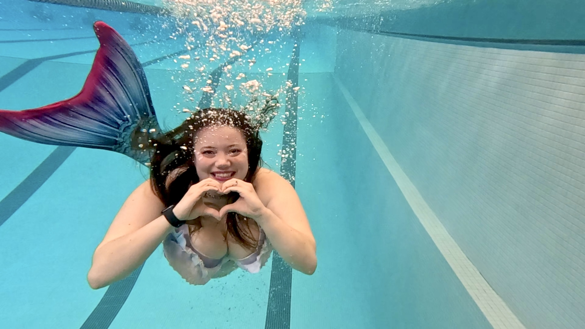 Young woman with a mermaid tail swimming underwater, smiling and making a heart shape with her hands.