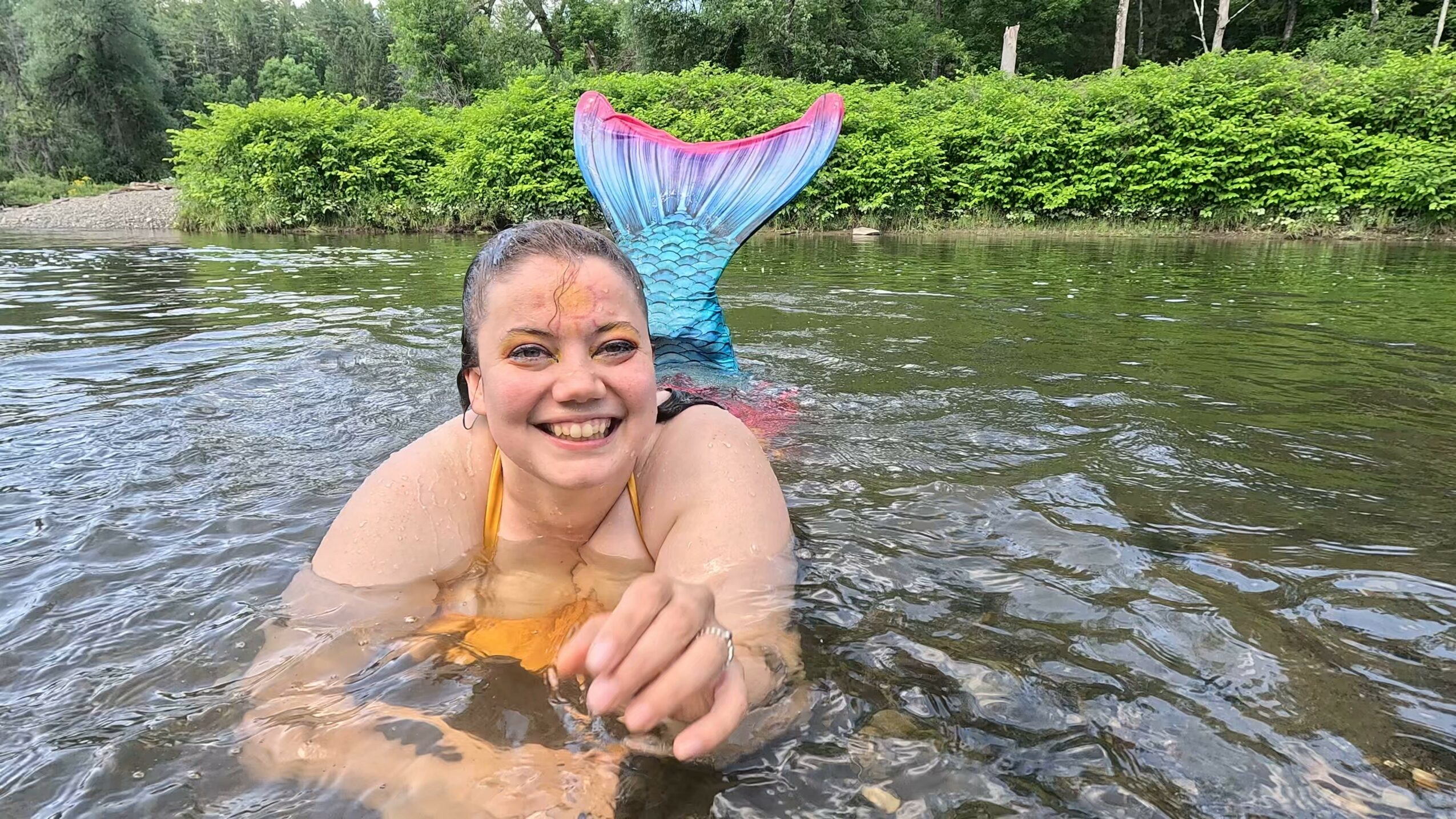 Person with a mermaid tail swimming in water, smiling, with greenery in the background.