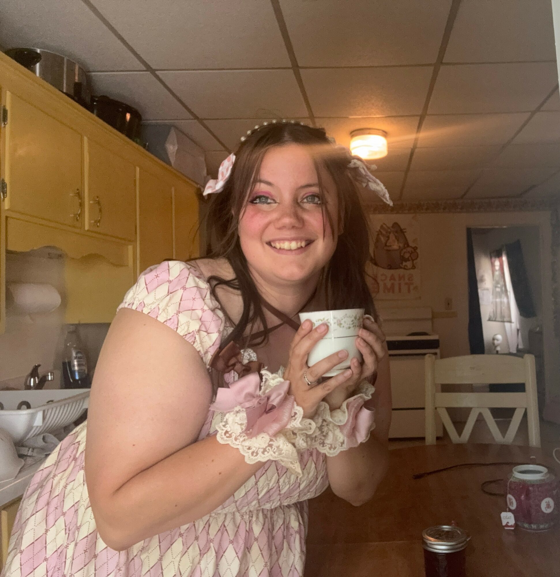 Smiling young woman holding a cup, wearing a pink and white dress with lace details, in a kitchen setting.
