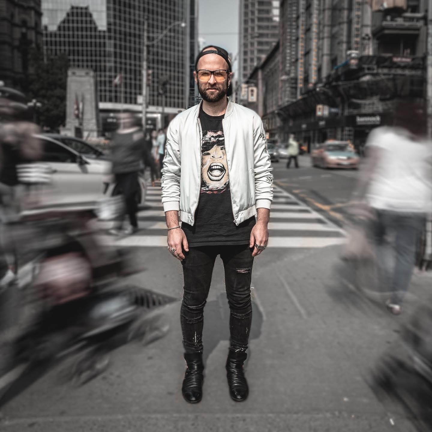 Man standing on city street crosswalk with blurred moving cars and pedestrians in background.