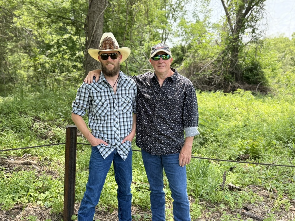 Two men standing outdoors in a green, wooded area, posing for a photo, one wearing a cowboy hat and sunglasses.