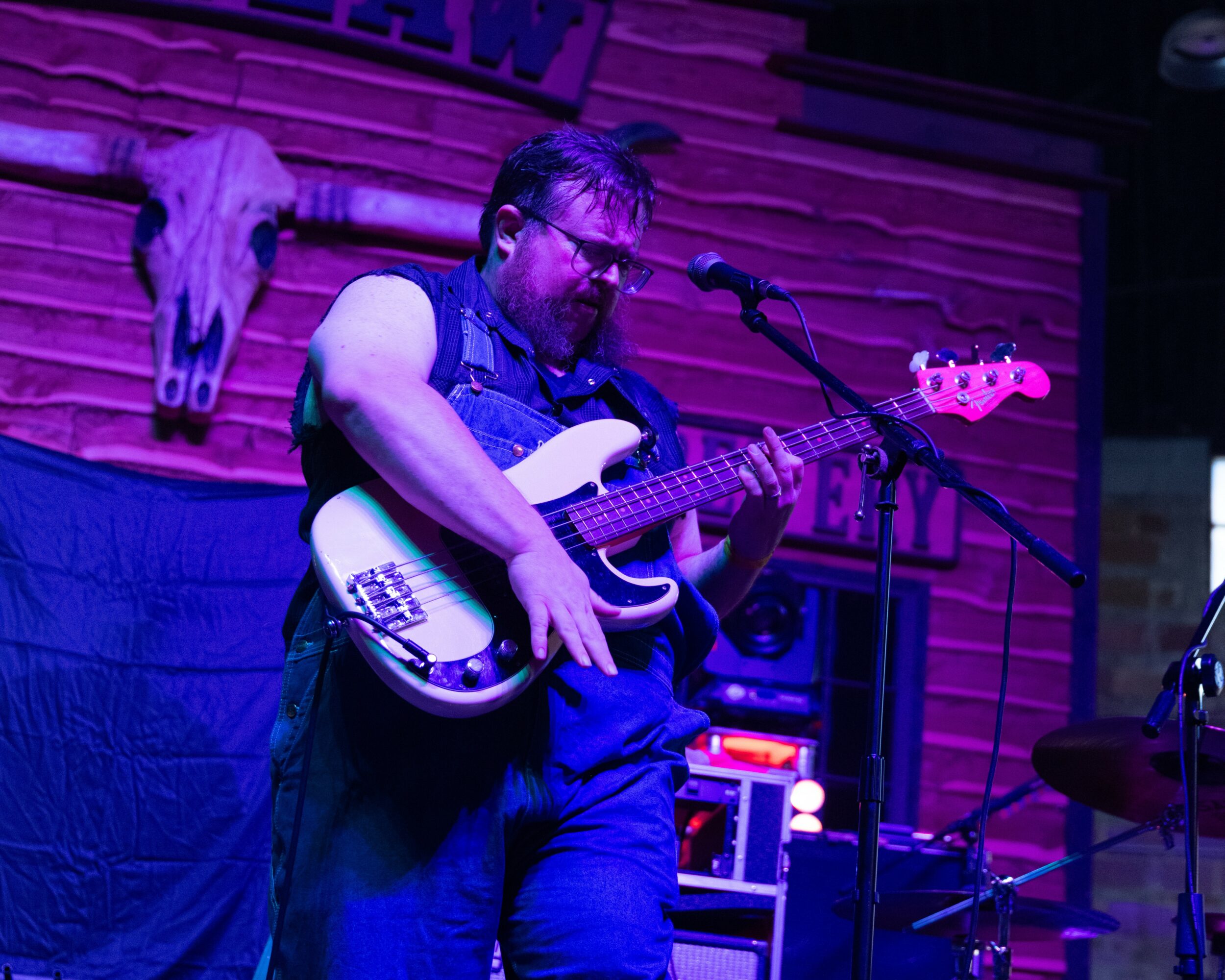 Person playing bass guitar on stage with wooden wall and animal skull in background.