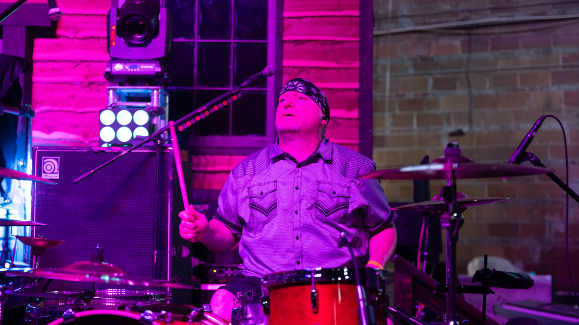 Person playing drums on stage with colorful lighting and equipment, brick wall background, wearing glasses and a bandana.