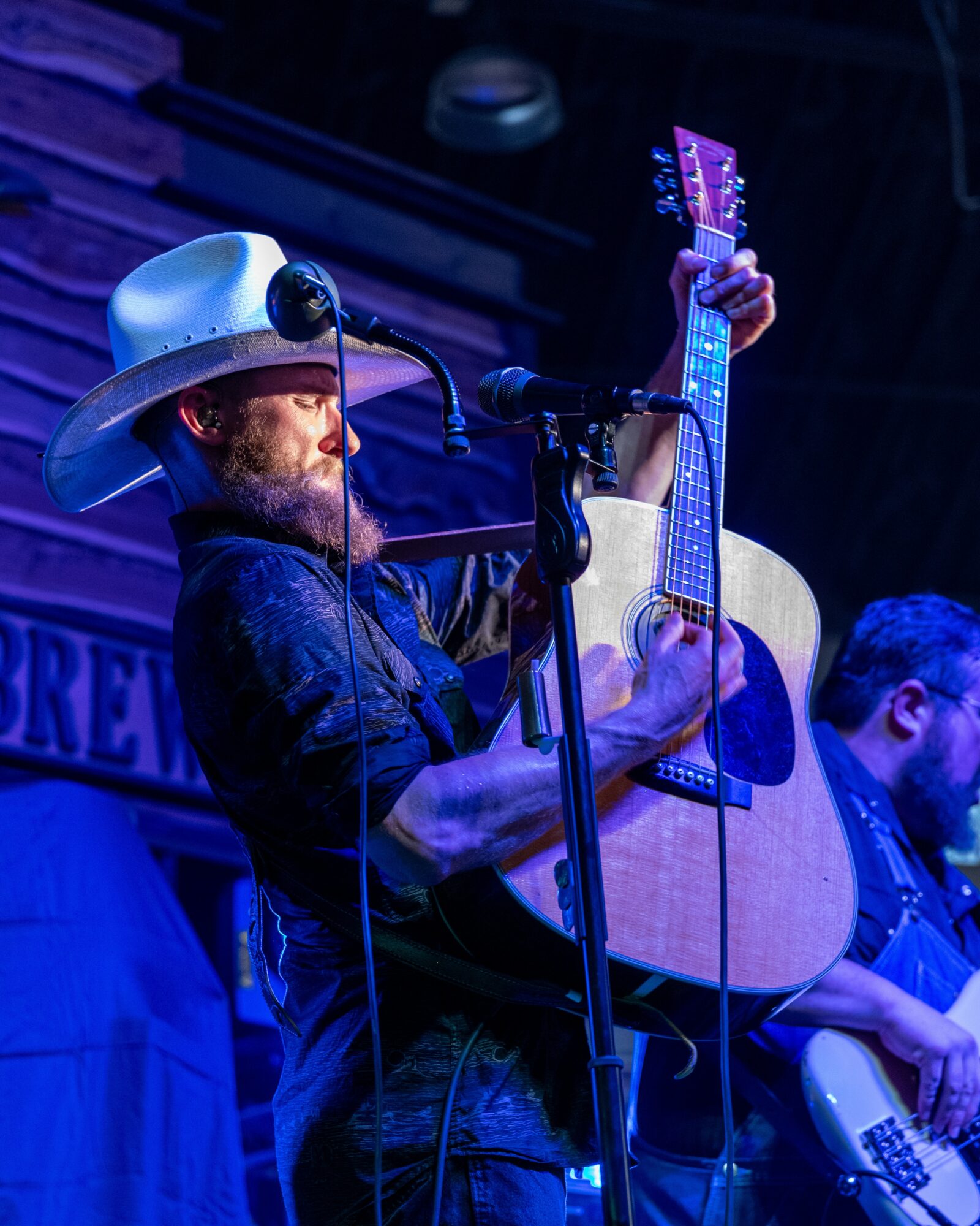 Musician with a cowboy hat playing an acoustic guitar on stage, with another musician in the background, under blue lighting.