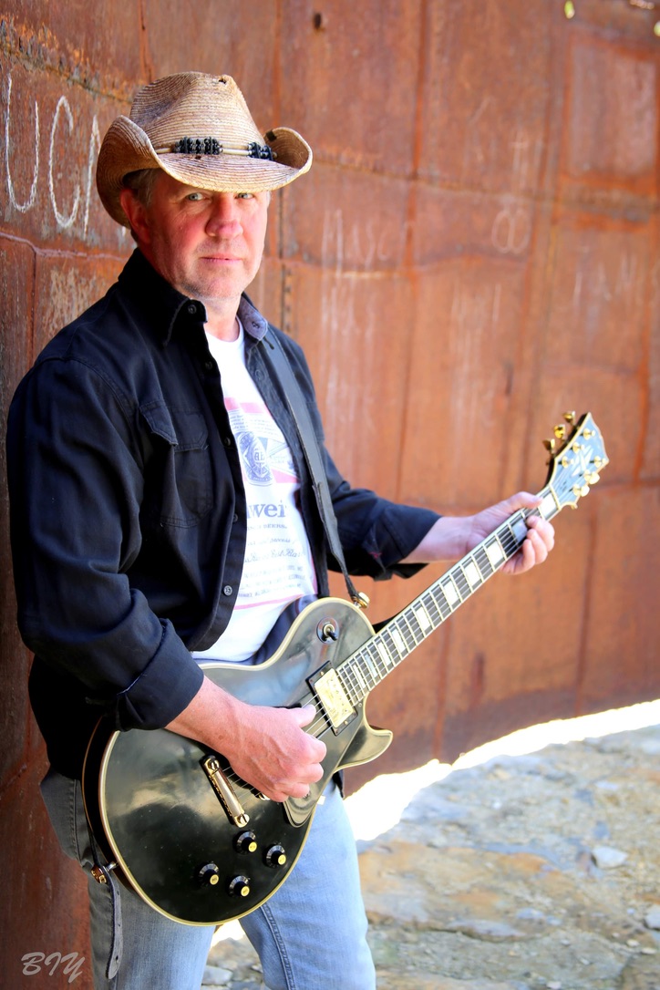 Man wearing a cowboy hat and black jacket playing a black electric guitar outdoors against a rust-colored wall.