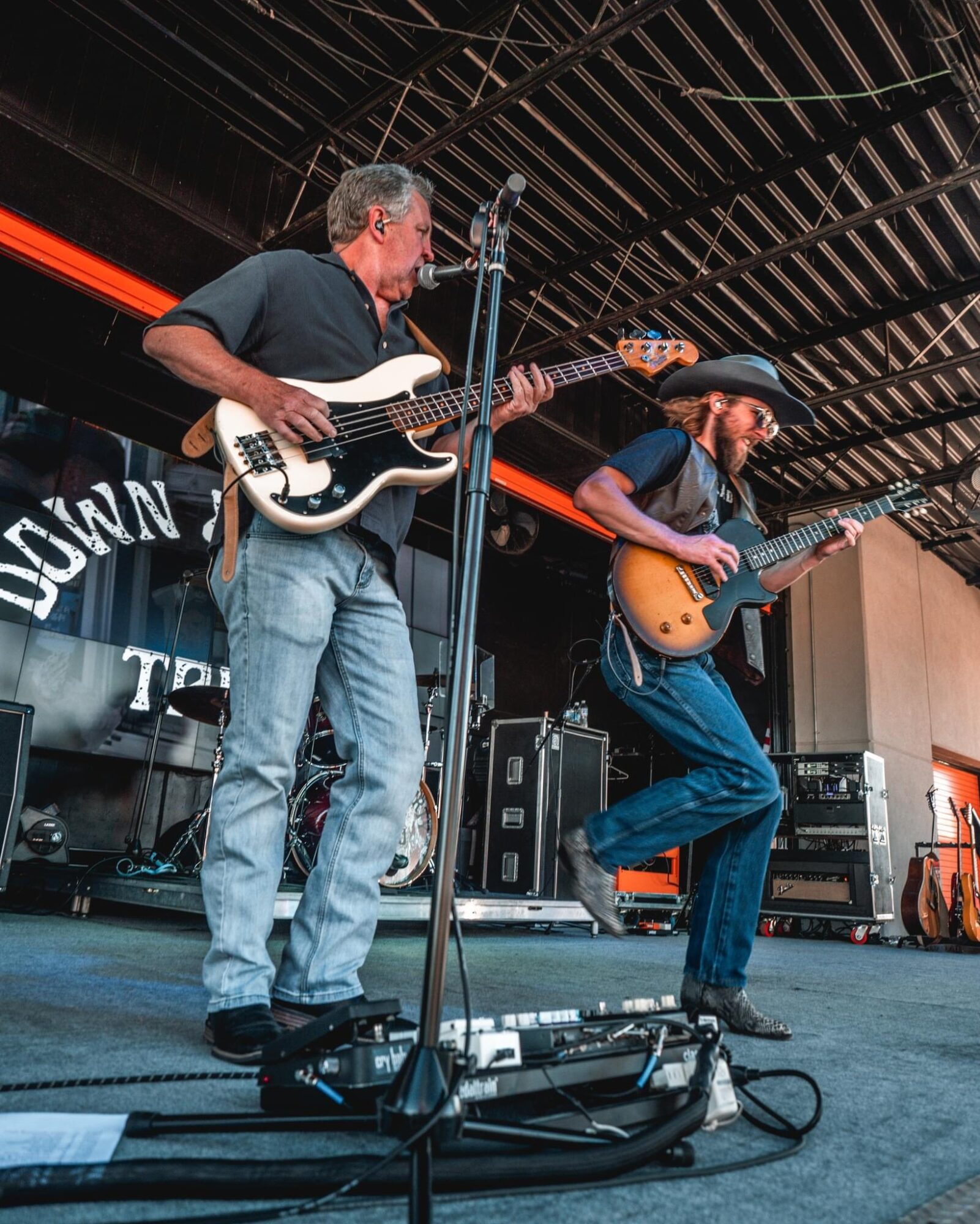 Two musicians perform on stage, one playing bass guitar and the other playing electric guitar, with stage equipment and a backdrop.