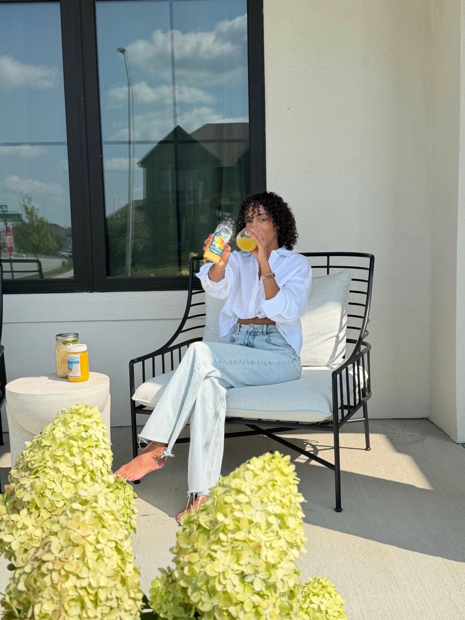 Person sitting on a black chair holding a water bottle, with a white wall and window behind, and yellow flowers in foreground.
