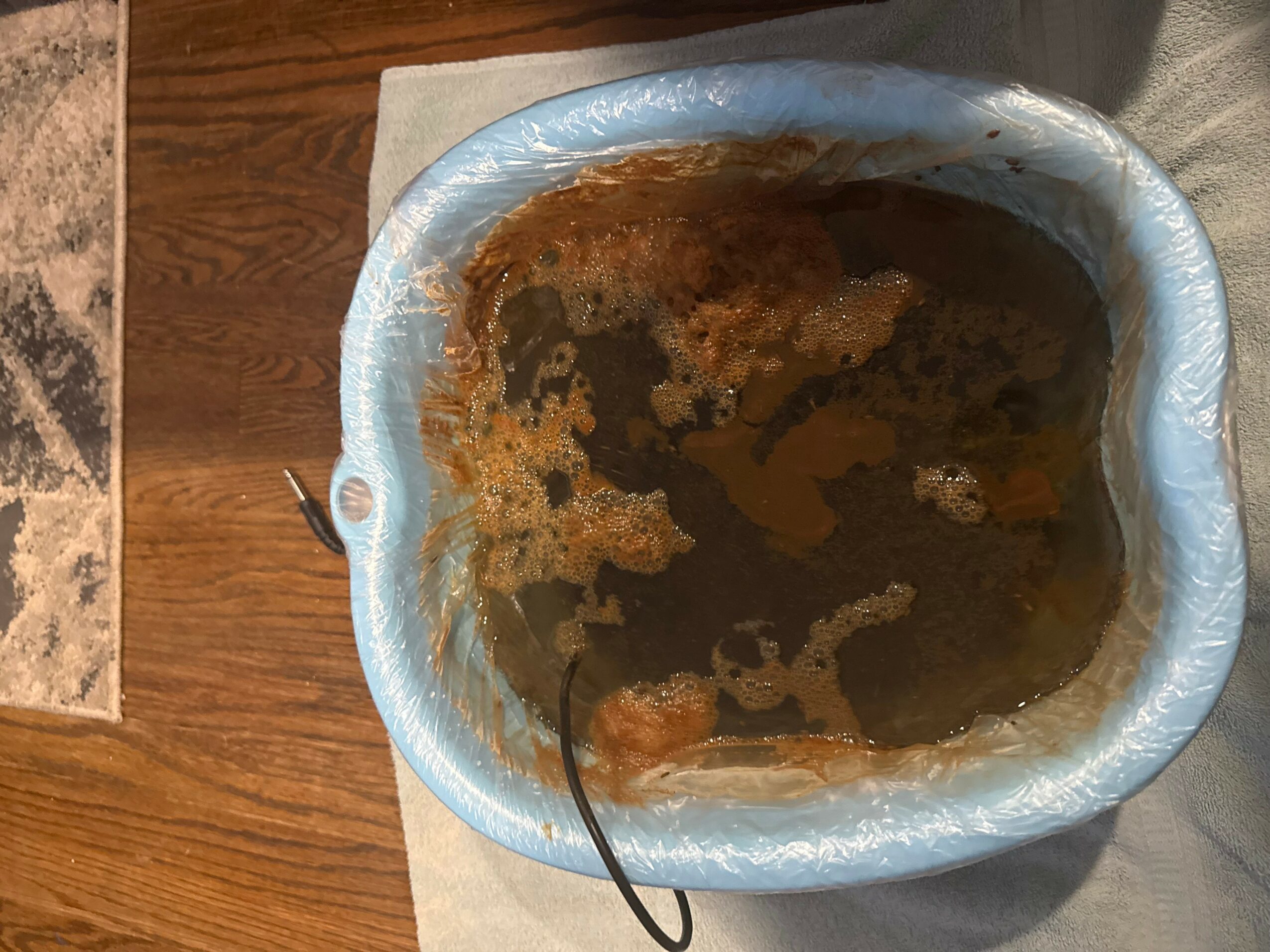 A bowl with a blue rim filled with dark, murky liquid and floating debris, placed on a white cloth on a wooden surface.