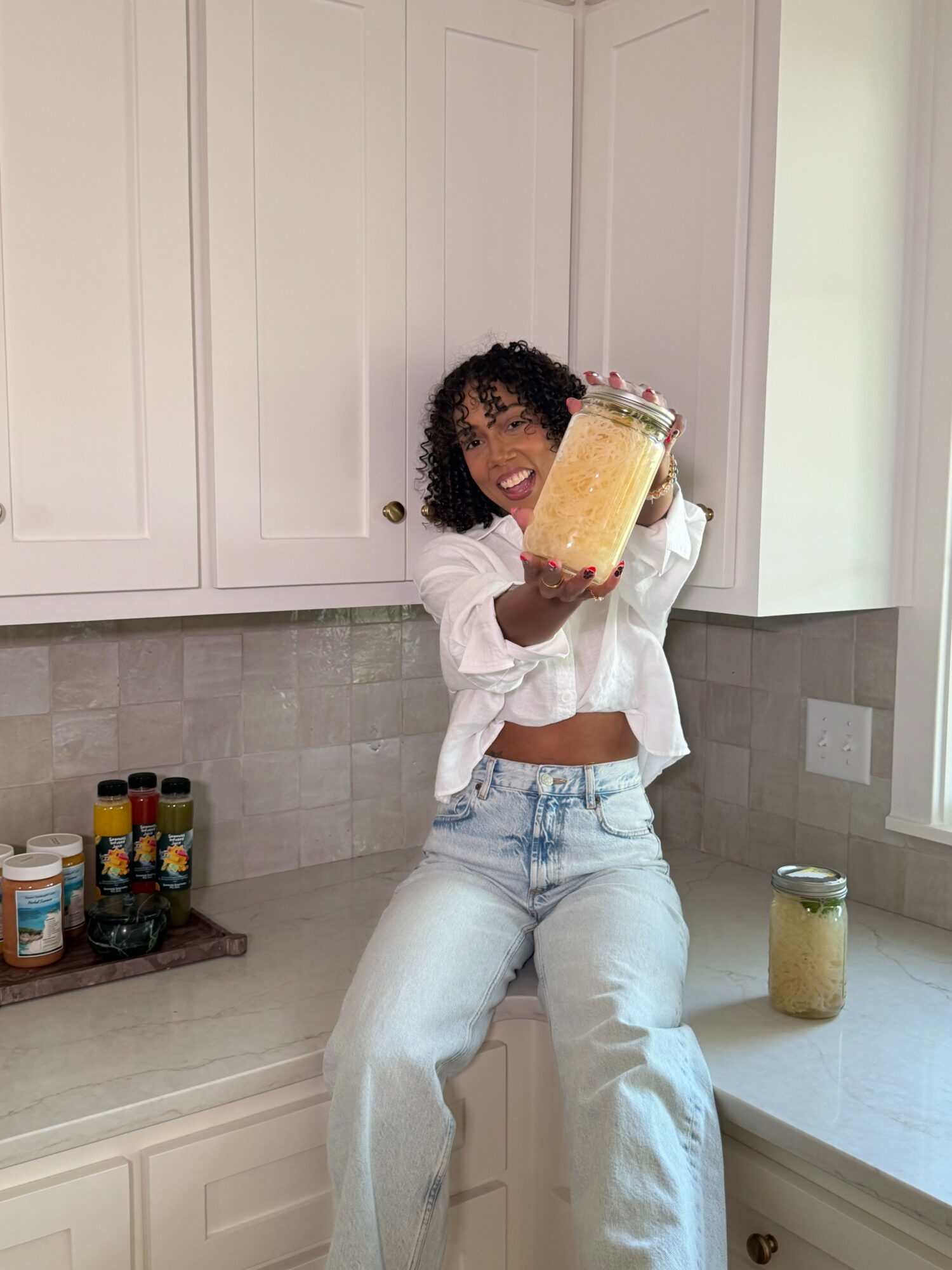 Person with curly hair holding a yellow drink in a glass, standing in a kitchen with white cabinets and a tiled floor.