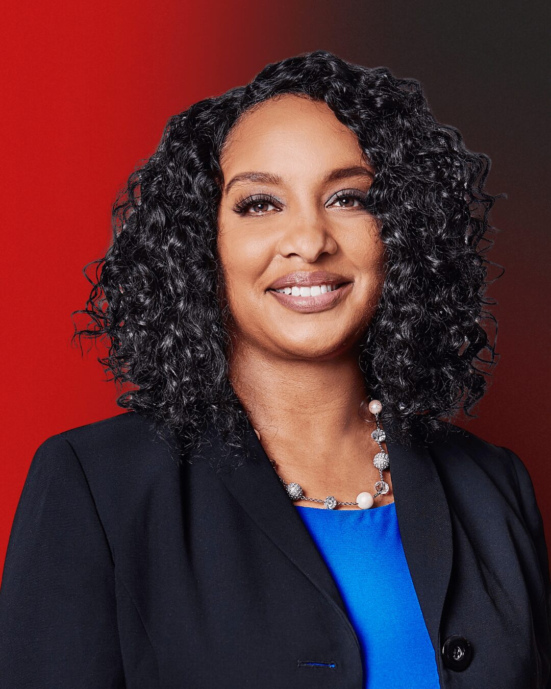 Woman with curly hair wearing a black blazer and blue top, smiling, against a red and dark background.