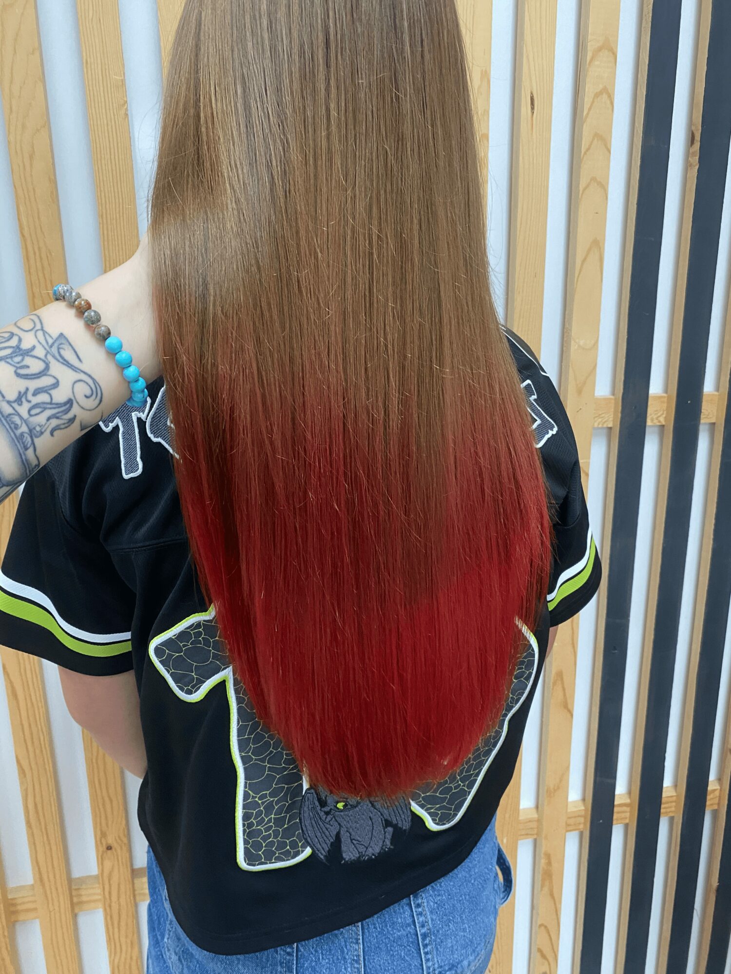 Person with long, straight hair dyed red at the ends, standing against a wooden slat background.