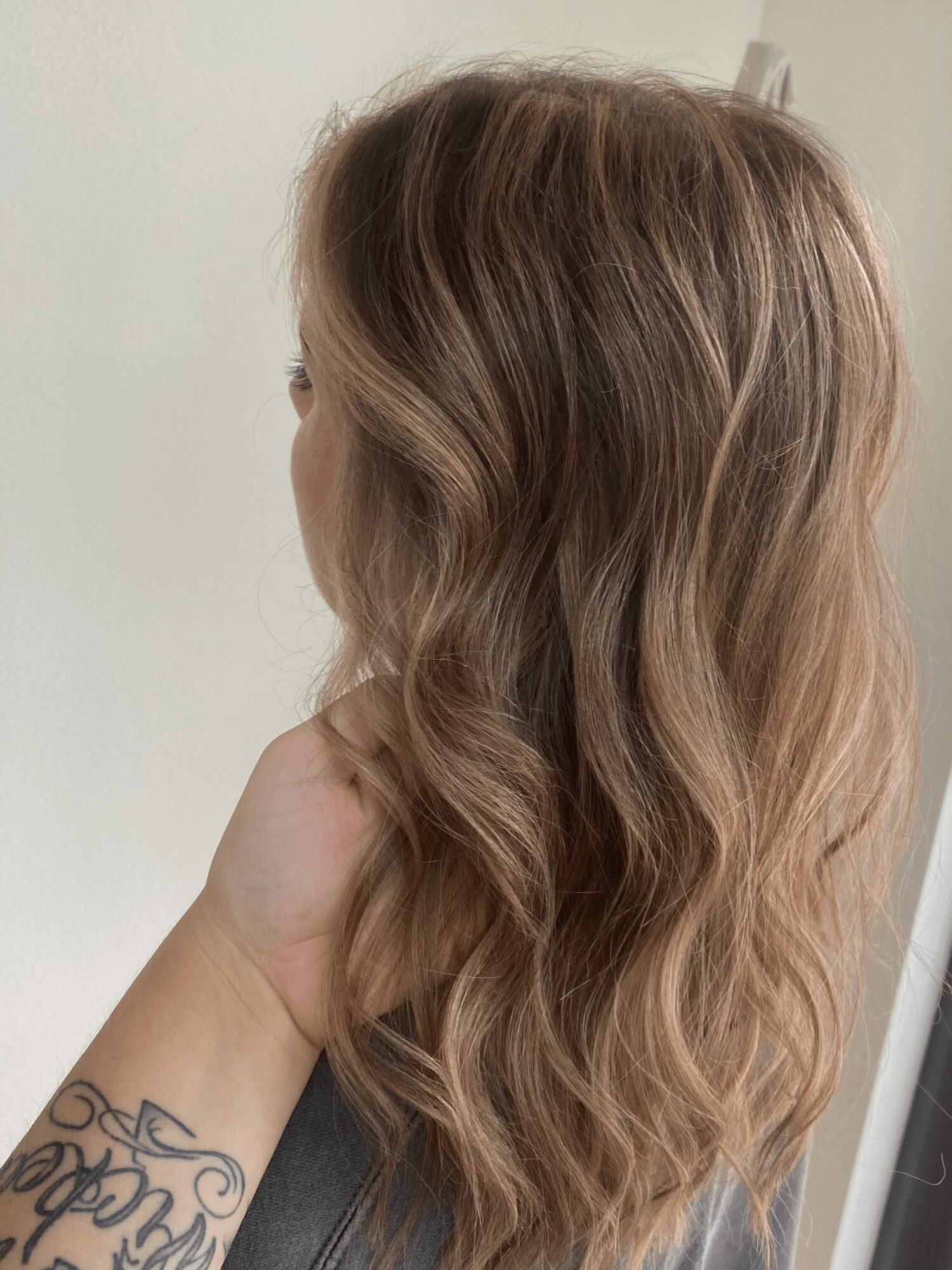 Person with long, wavy, light brown hair viewed from the back, hand touching hair, neutral background.