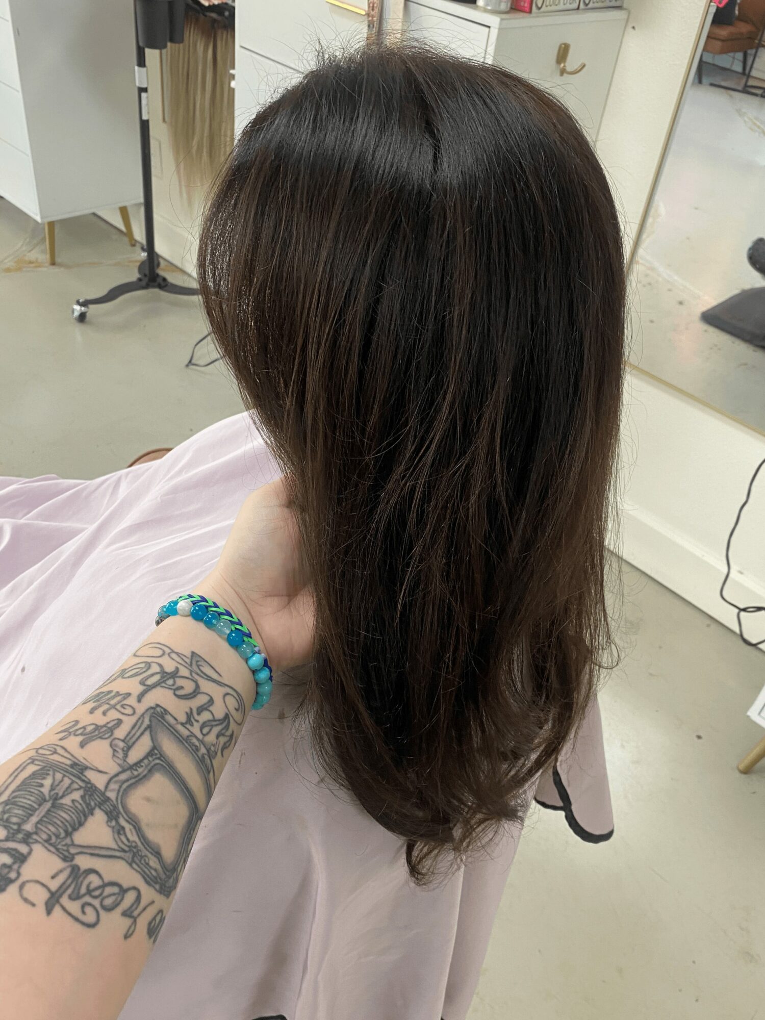 Person with long, dark brown hair sitting in a salon chair, hand with tattoo and bracelet visible, background of salon interior.