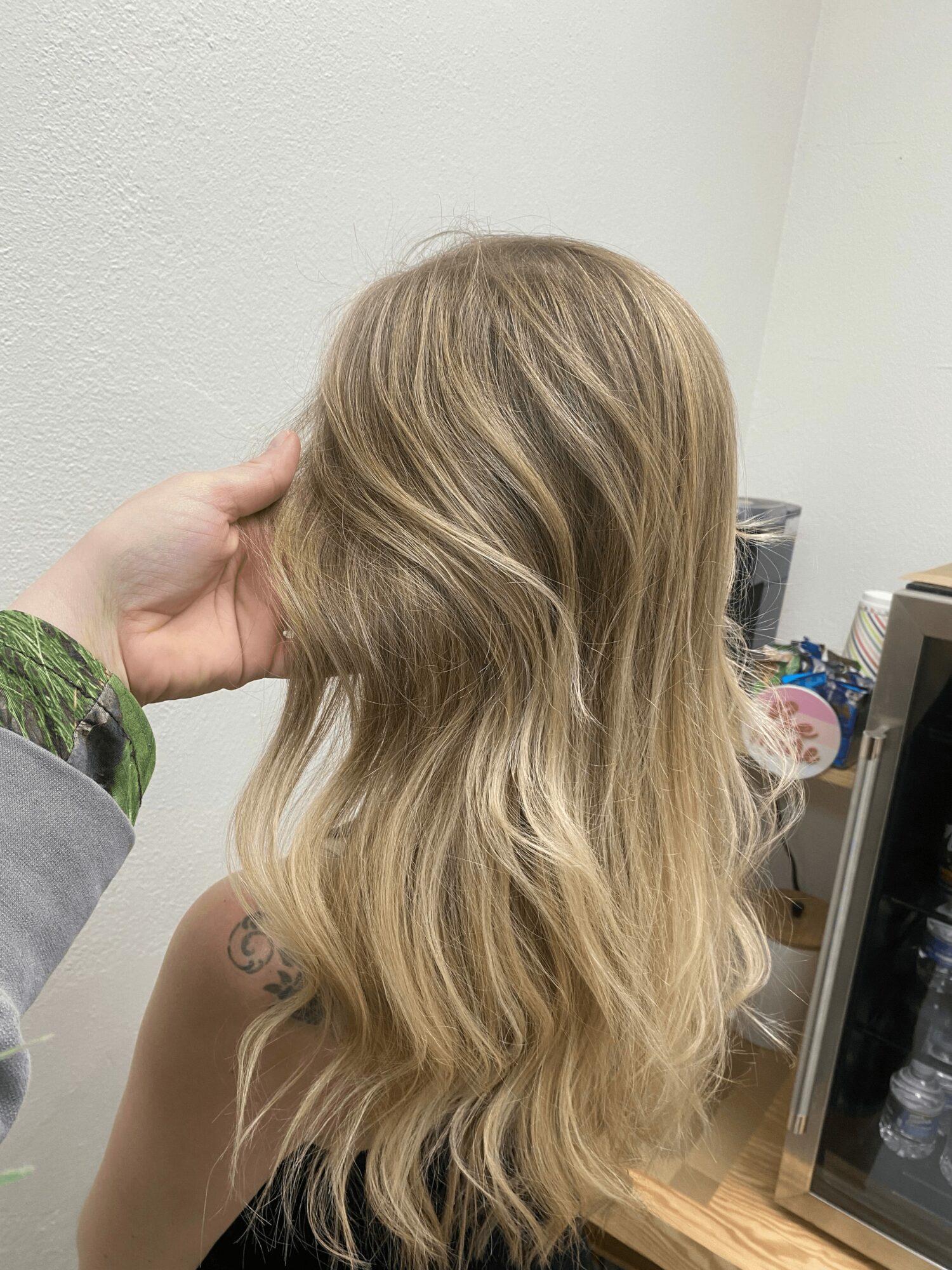 Person with long, wavy blonde hair, hand touching hair, in a room with a white wall and a desk.