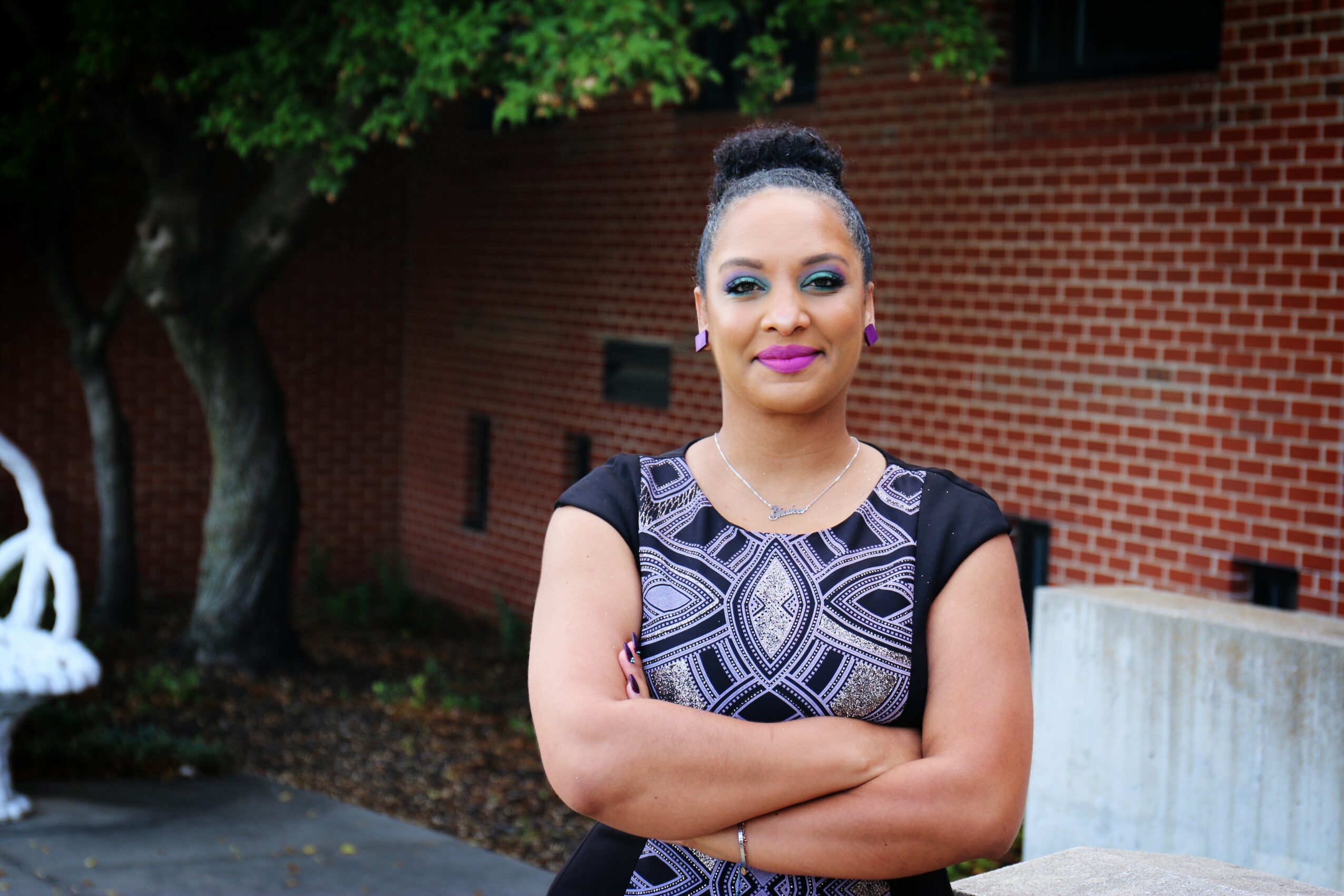 Woman with arms crossed, standing outdoors in front of a brick building and tree, smiling confidently.