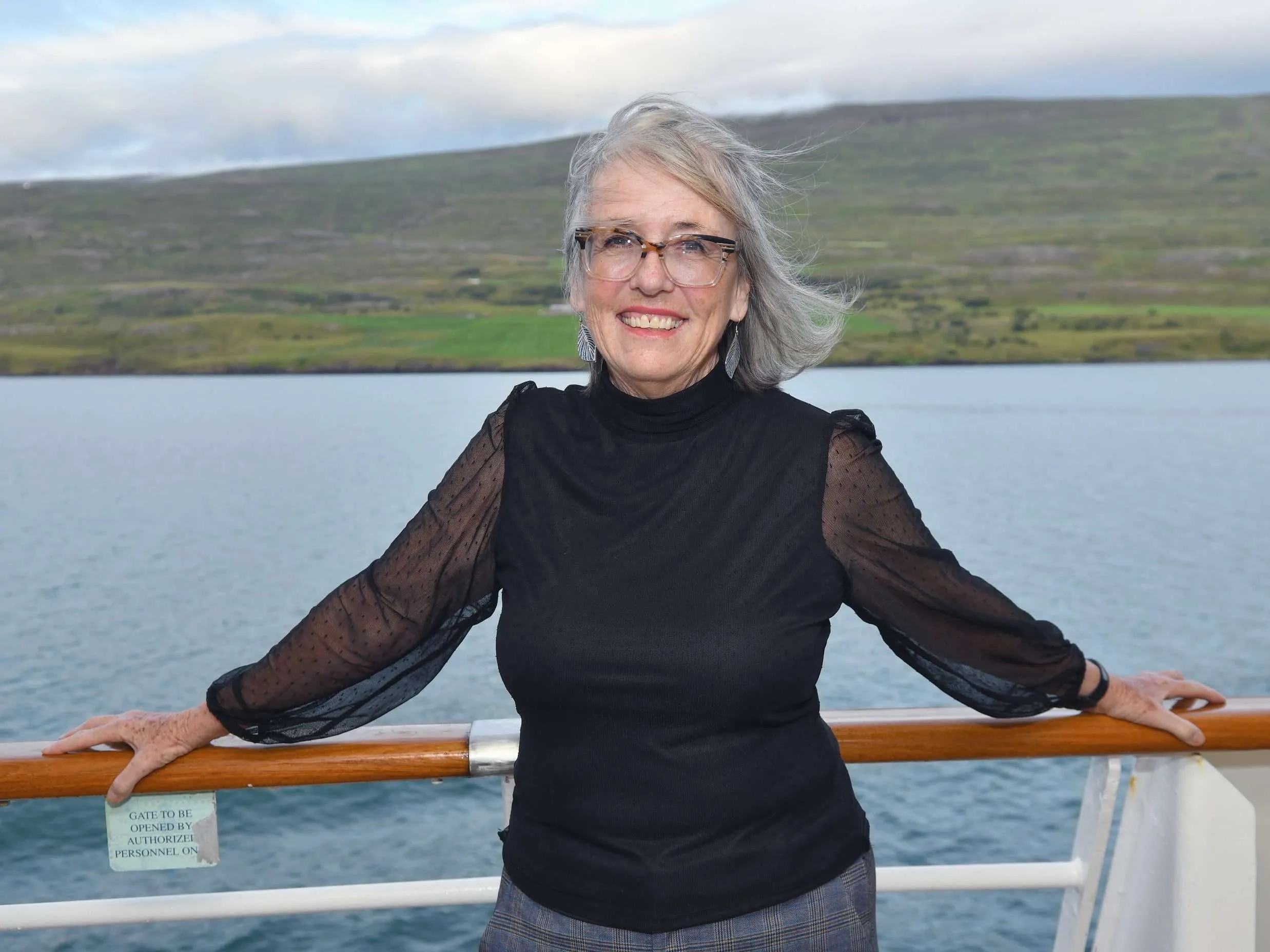 Smiling woman with gray hair and glasses stands outdoors near water, with a hill in the background, holding a railing.
