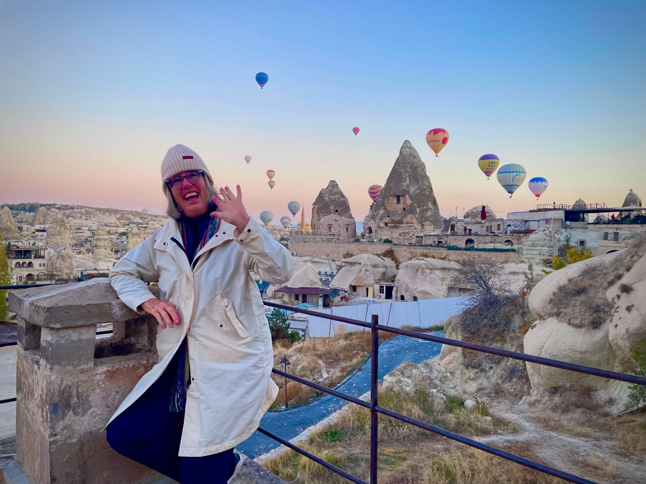 Person in white jacket and hat waving, hot air balloons in sky over rocky landscape and buildings, early morning or late afternoon light.