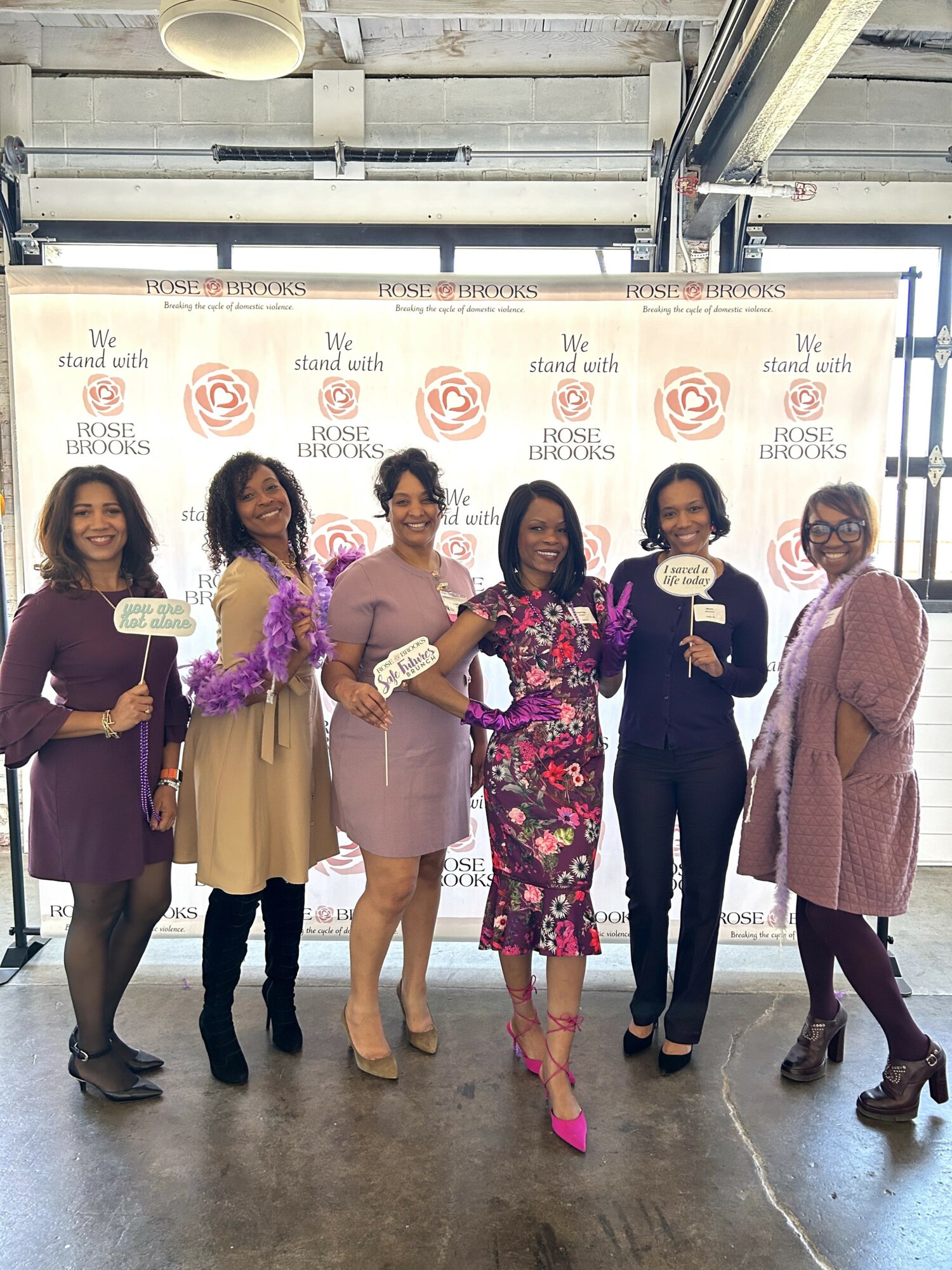 Seven women standing in front of a backdrop with logos, holding signs, smiling, in an indoor setting.