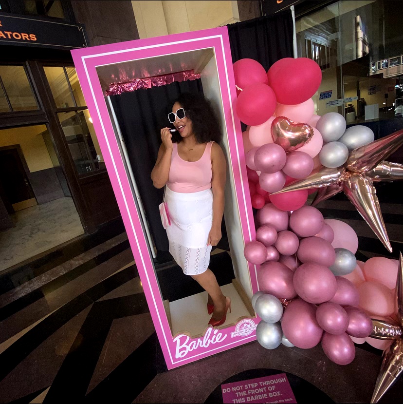Woman in pink top and white skirt taking a selfie in a pink Barbie-themed mirror surrounded by pink, silver, and star-shaped balloons.