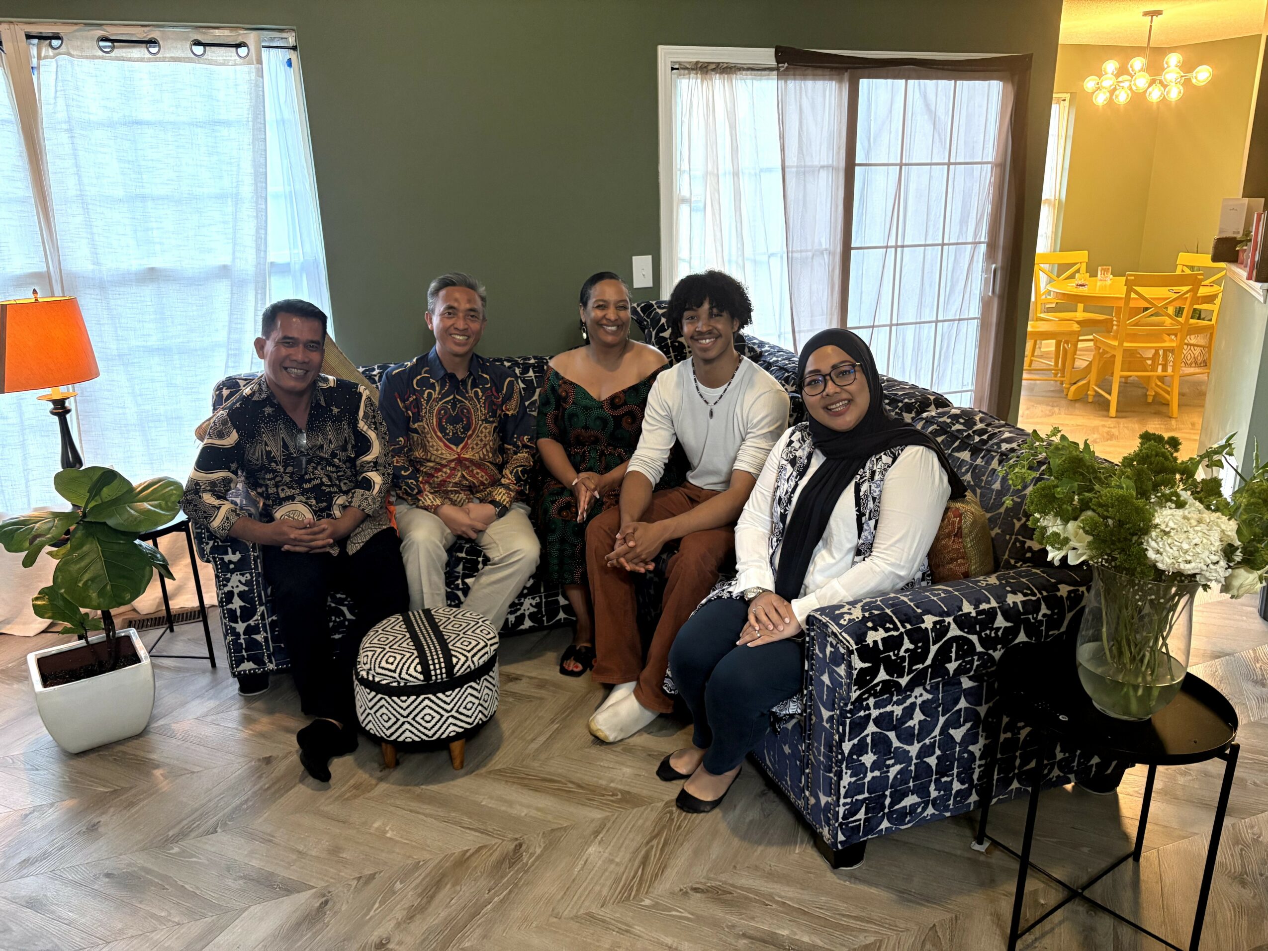 Five people sitting on sofas and chairs in a living room, smiling, with plants and windows in the background.