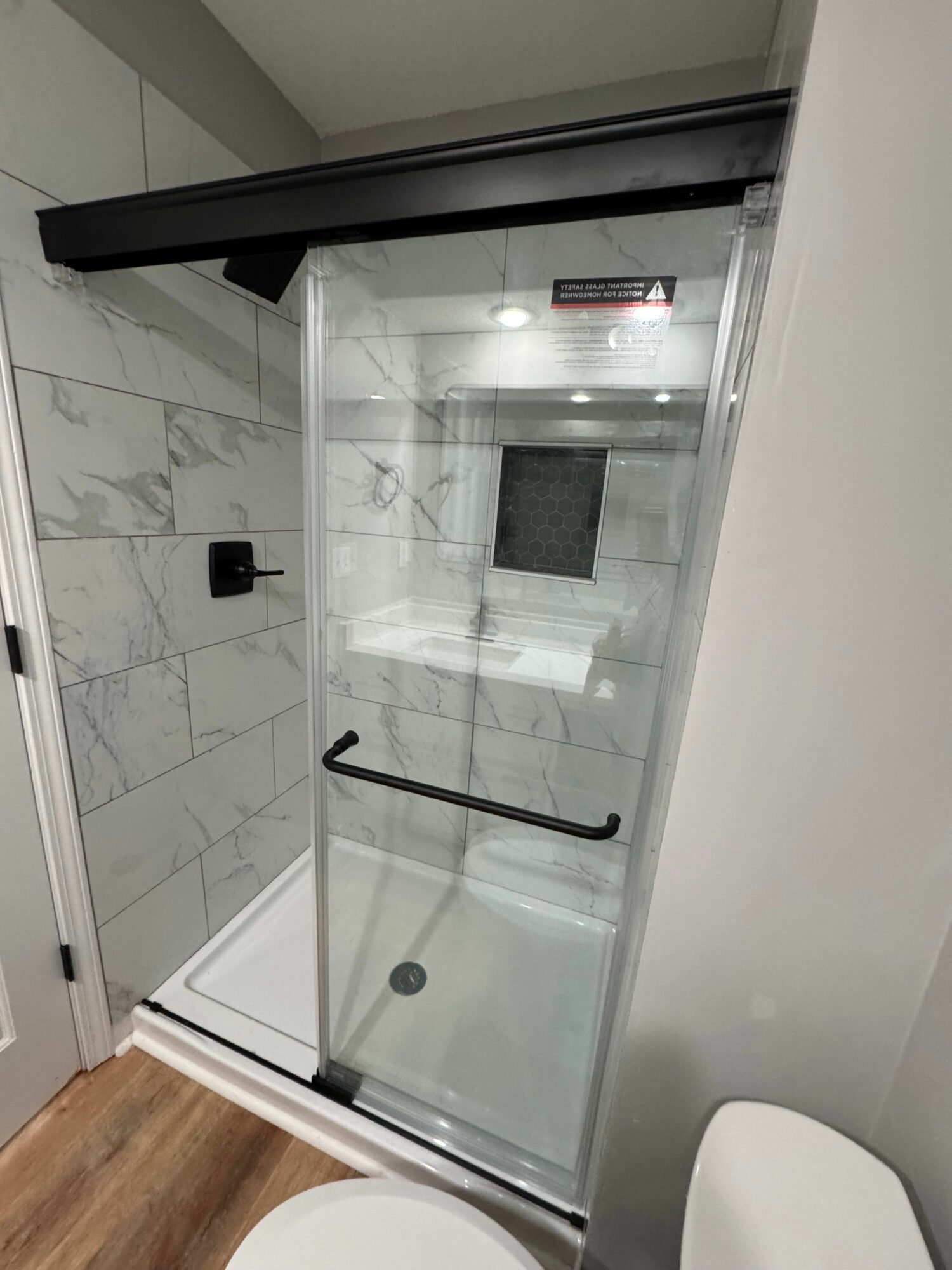 Shower area with glass door, marble floor, and a black handle, viewed from outside the shower.