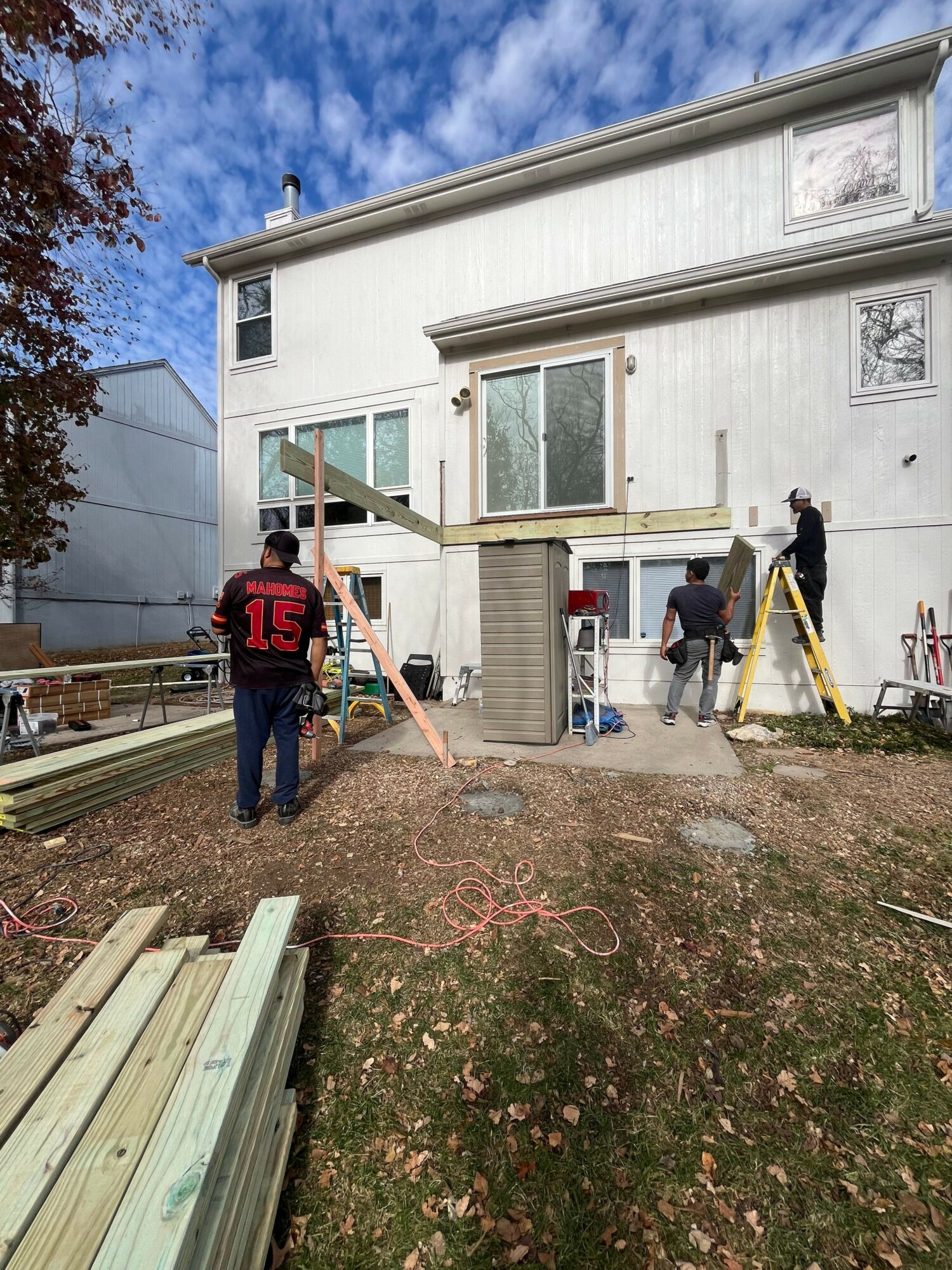 People working on a house construction or renovation outside, with building materials and tools around, under a partly cloudy sky.